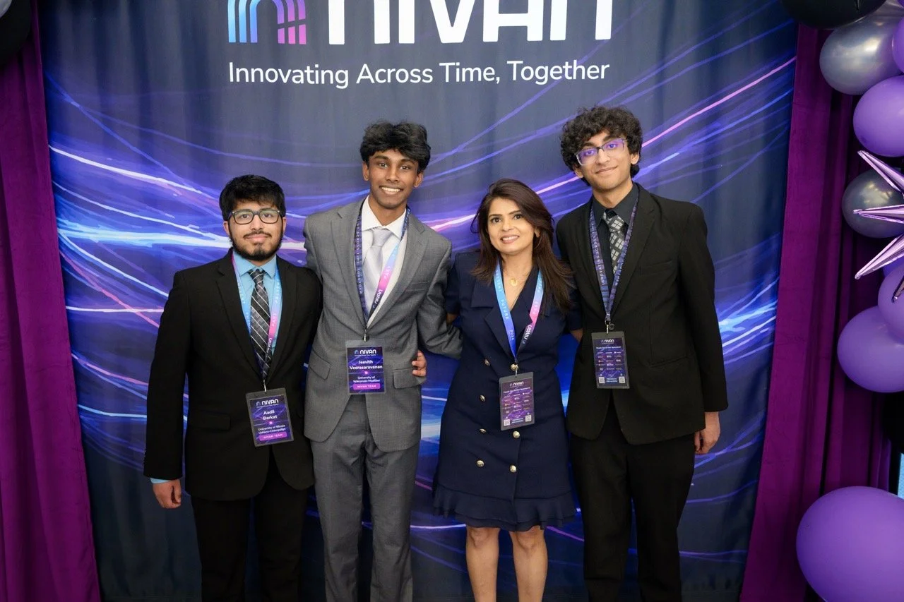 Four people in formal attire standing together , smiling for the camera in front of a backdrop that reads 'NIVAN' with the slogan 'Innovating Across Time, Together.' The backdrop has a dark background with blue and purple streaks, and there are purpl