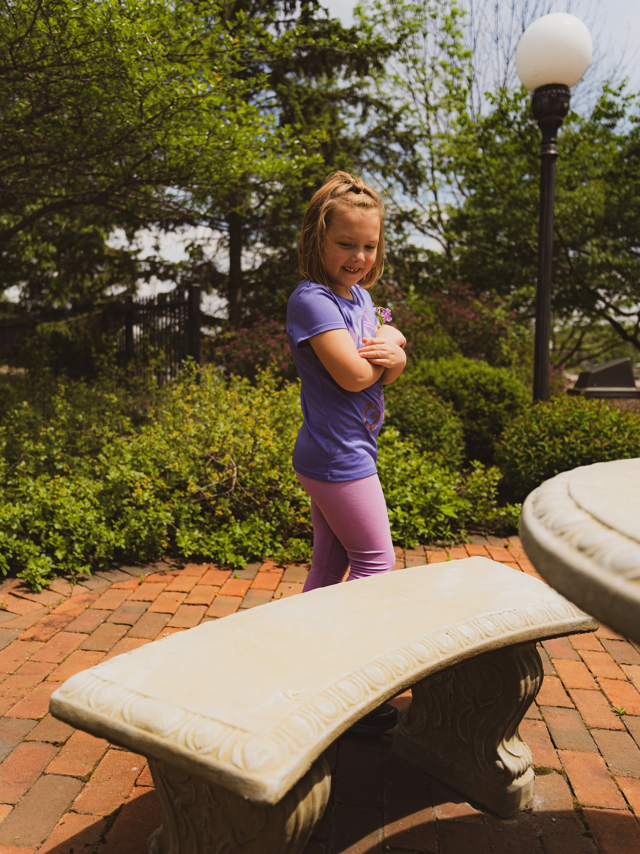 A young girl wearing a purple shirt and pink leggings stands outdoors in a garden, smiling and holding a small bunch of flowers, with a stone bench in the foreground and trees and a lamppost in the background.