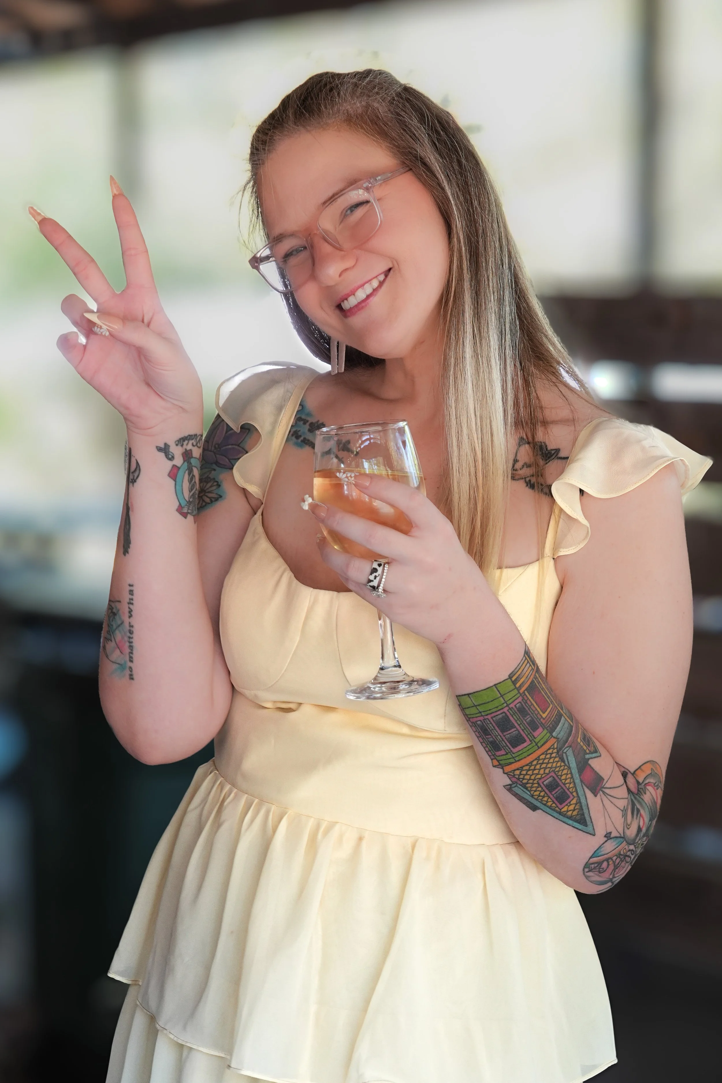 A woman smiling, holding a glass of white wine, peace sign with her right hand, wearing glasses, a yellow dress, and showing colorful tattoos on her arms and shoulders, indoors with a blurred background.