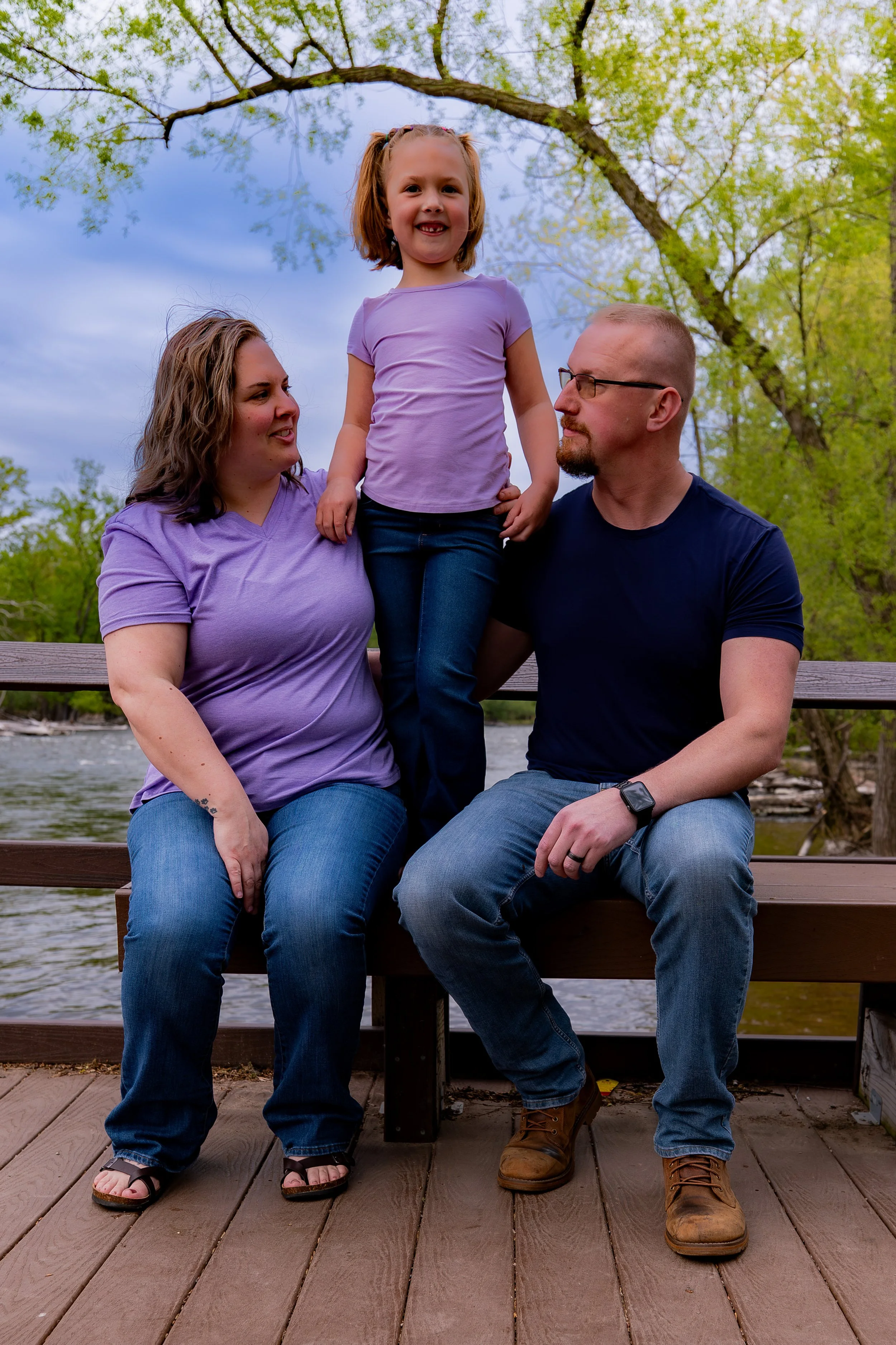 A family of three, a woman, a man, and a young girl, sitting on a wooden dock by a river, with green trees and a cloudy sky in the background. The girl is standing on the bench between the adults, smiling at the camera. The woman and man are looking 