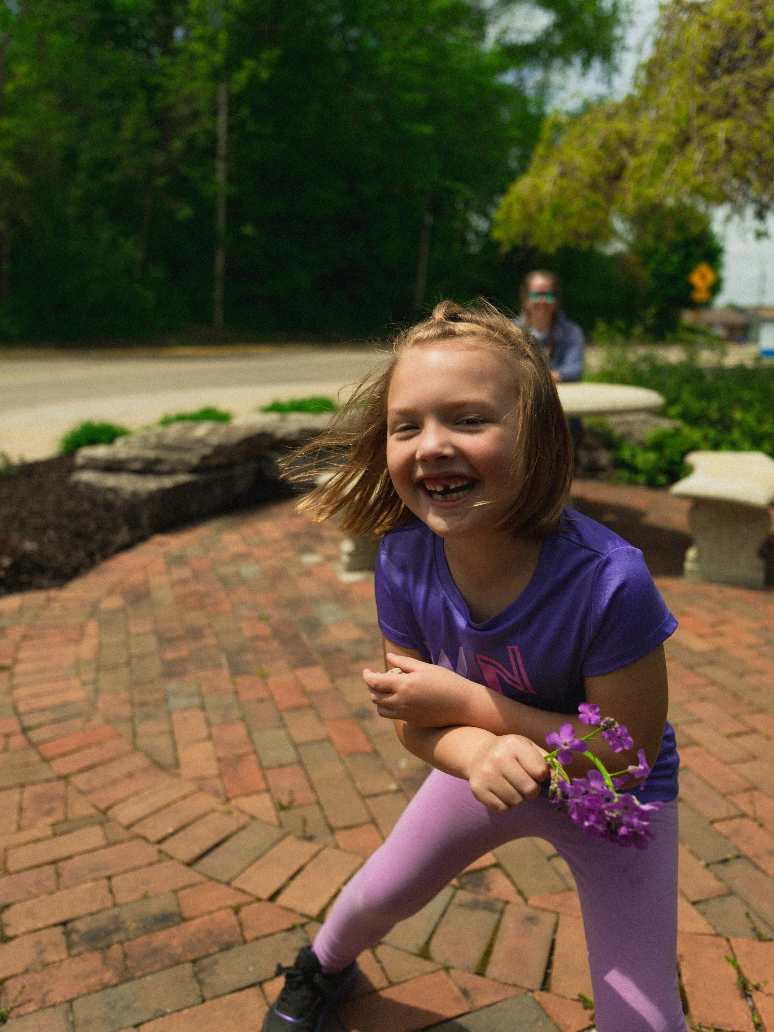 A young girl with red hair smiling and holding purple flowers outdoors on a brick-paved area, with a woman in the background near trees and greenery.