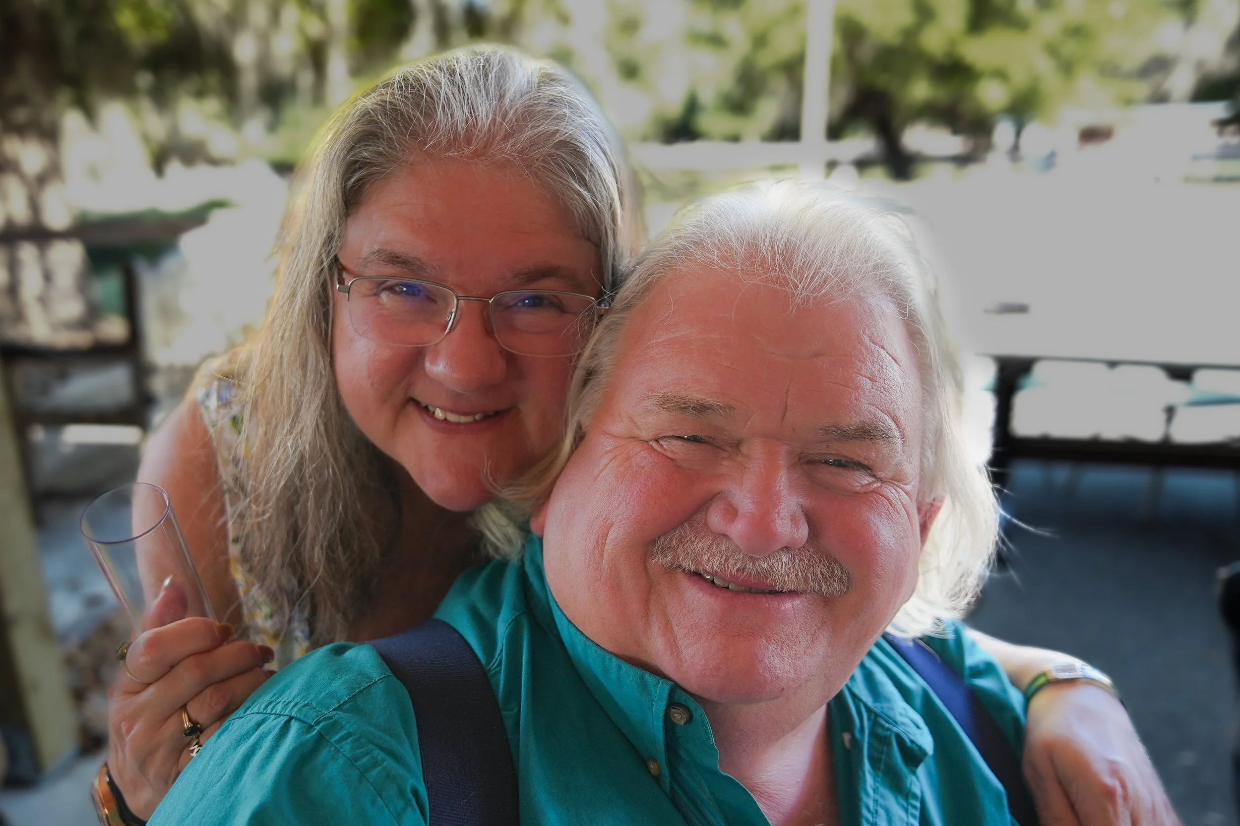 A smiling woman with long gray hair and glasses leaning over an older man's shoulder, both enjoying a moment together outdoors.