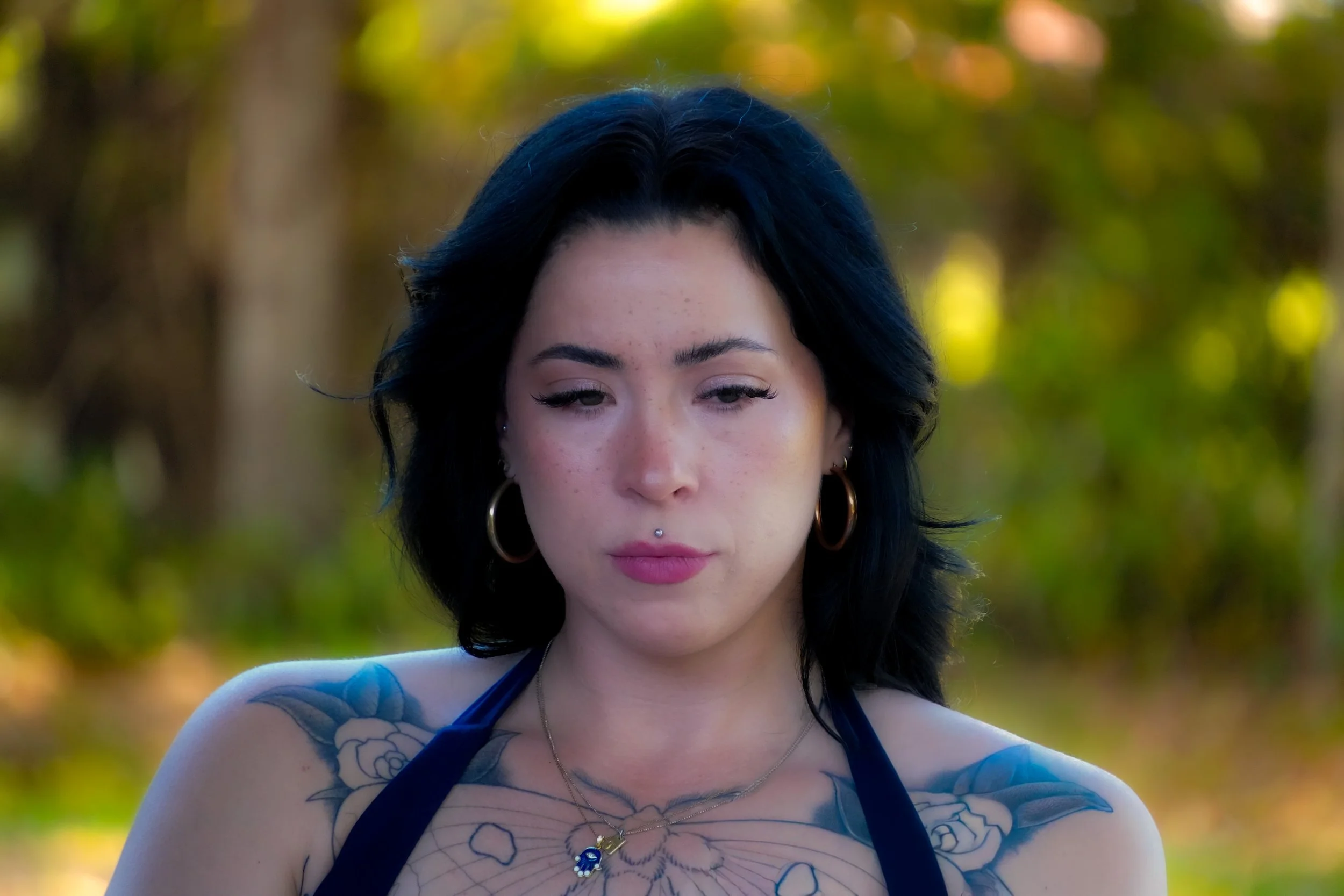 Portrait of a woman with dark hair, tattoos on her shoulders, and large hoop earrings, outdoors with a blurred background of trees and sunlight.