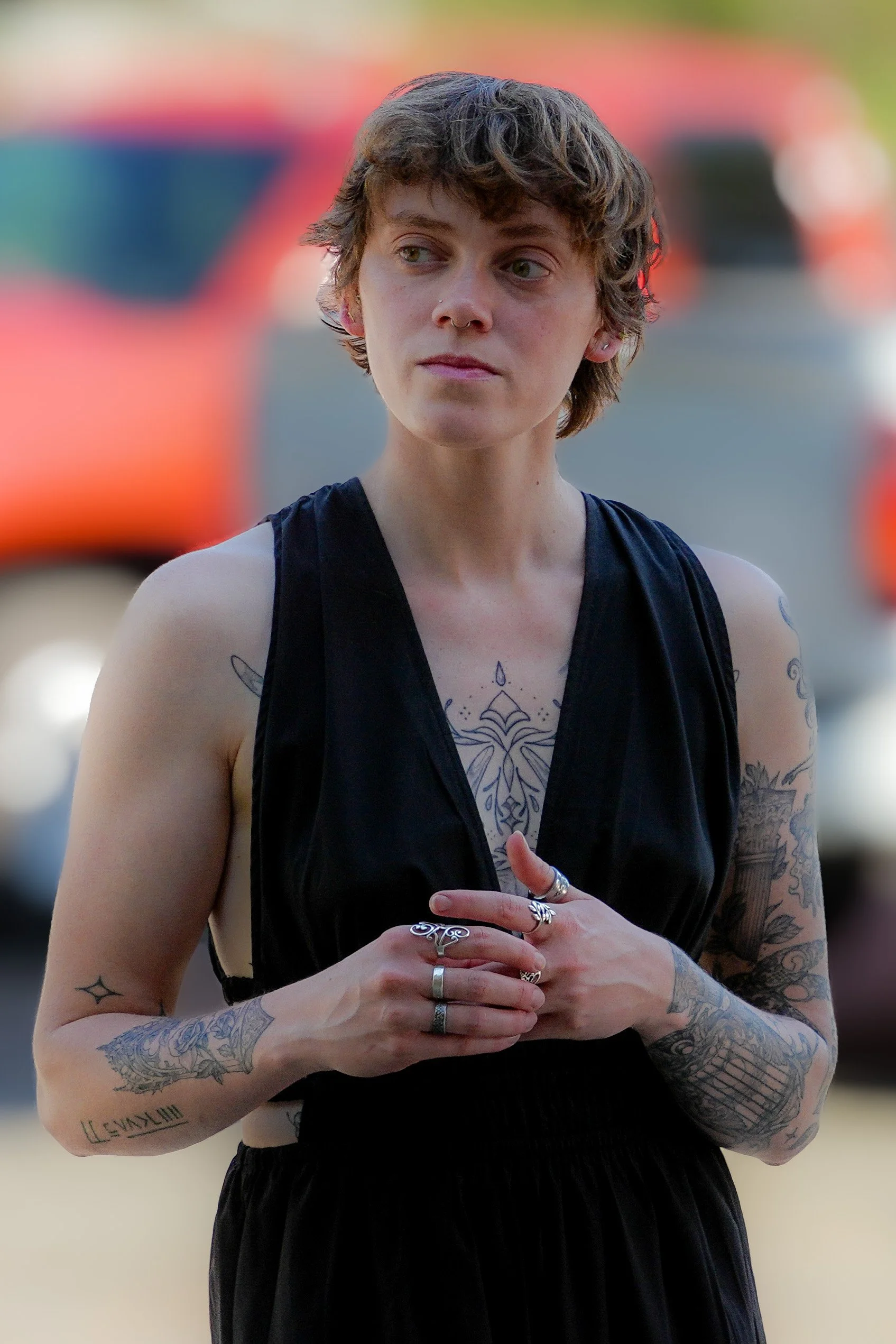 A young woman with short curly hair and tattoos on her arms and chest, dressed in a black sleeveless top, standing outdoors with a blurred background of people wearing orange and dark clothing.