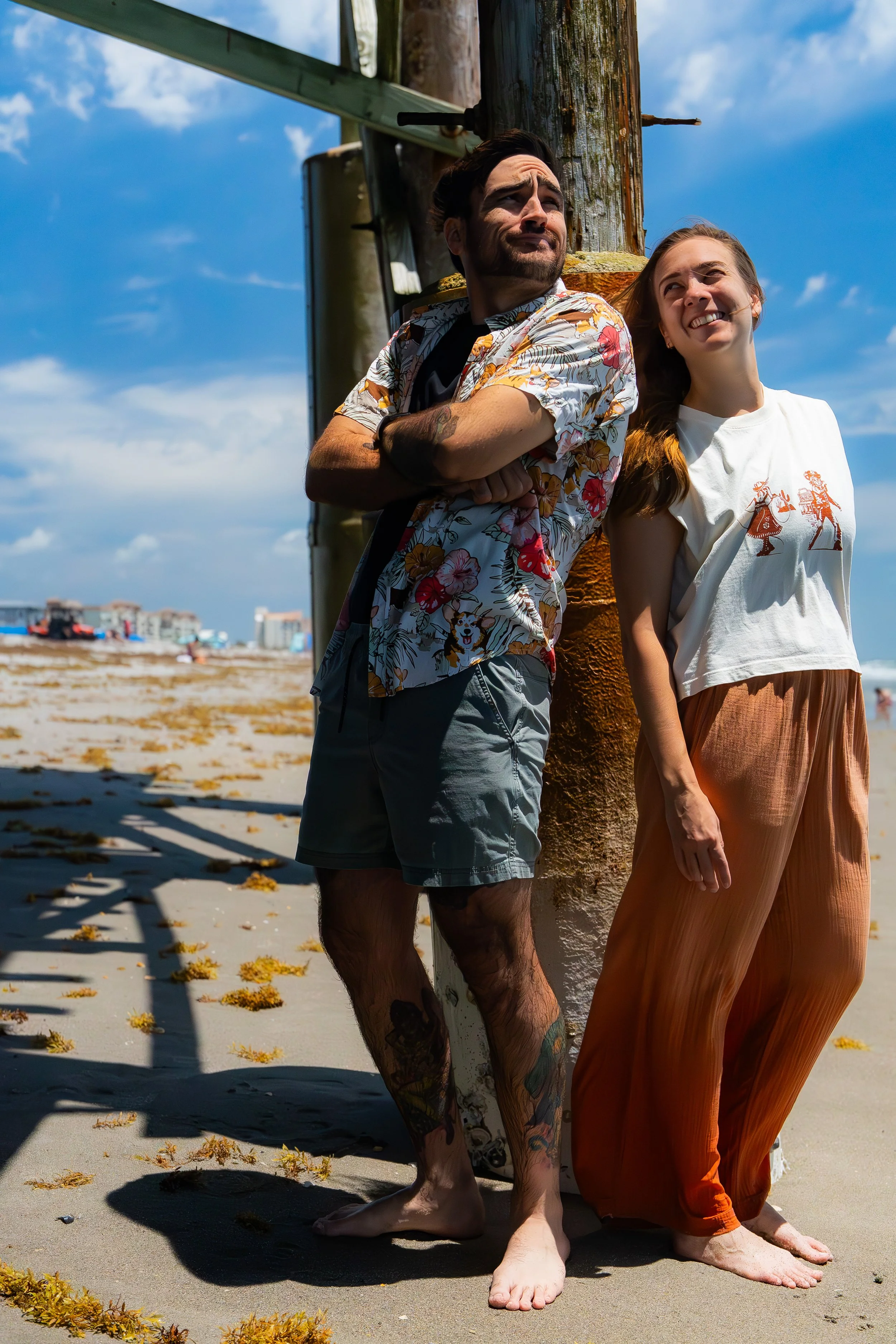 Two people standing on a beach, leaning against a wooden pole. The man has tattoos and is wearing a floral shirt, the woman is wearing a white t-shirt with a graphic and orange pants. The sky is blue with some clouds, and there are buildings and boat