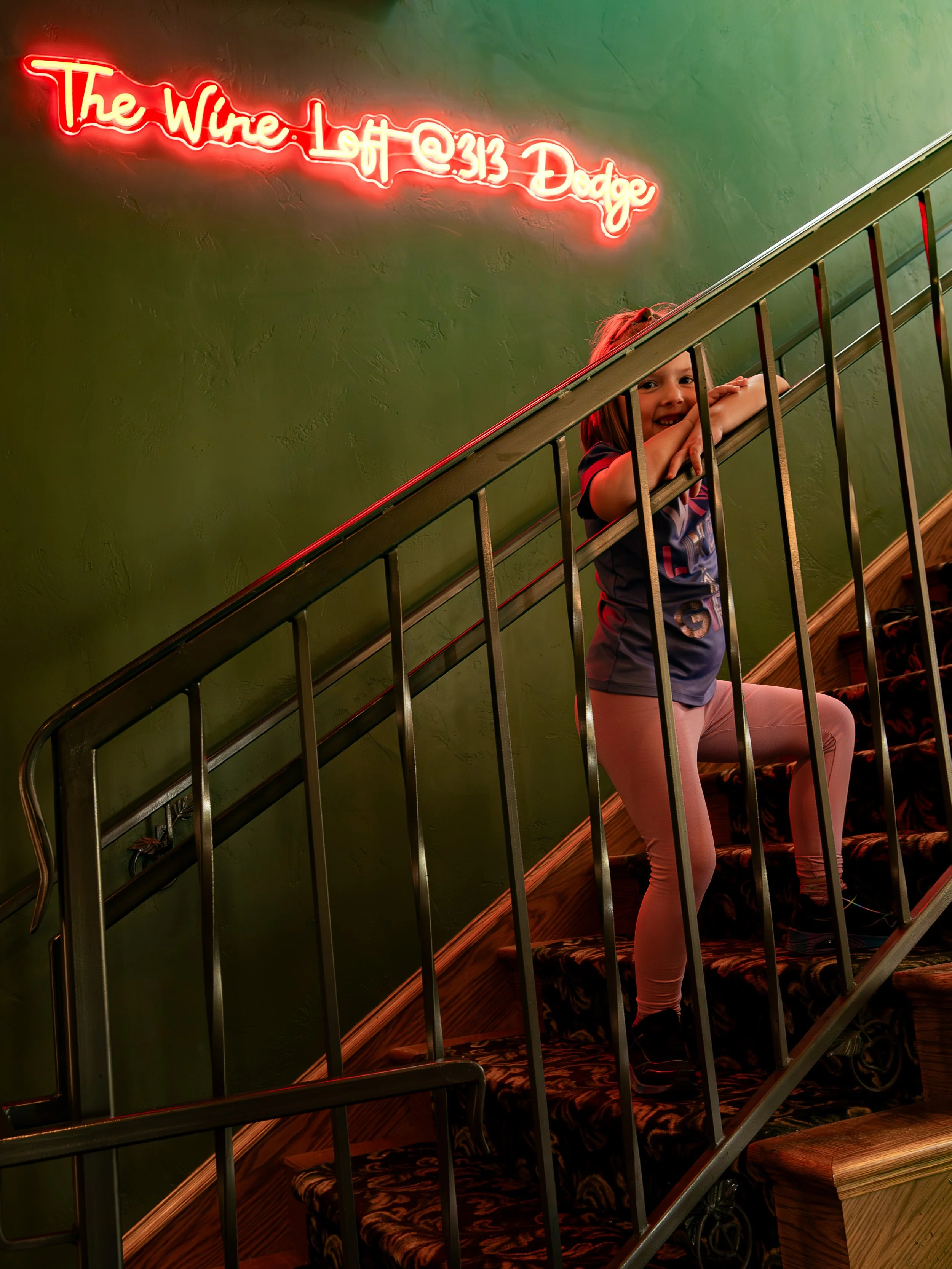 A young girl with red hair in a ponytail is standing on a staircase with metal railings, smiling and resting her head on her crossed arms. She is wearing pink pants and a black and red sports shirt. Behind her is a green textured wall with a red neon