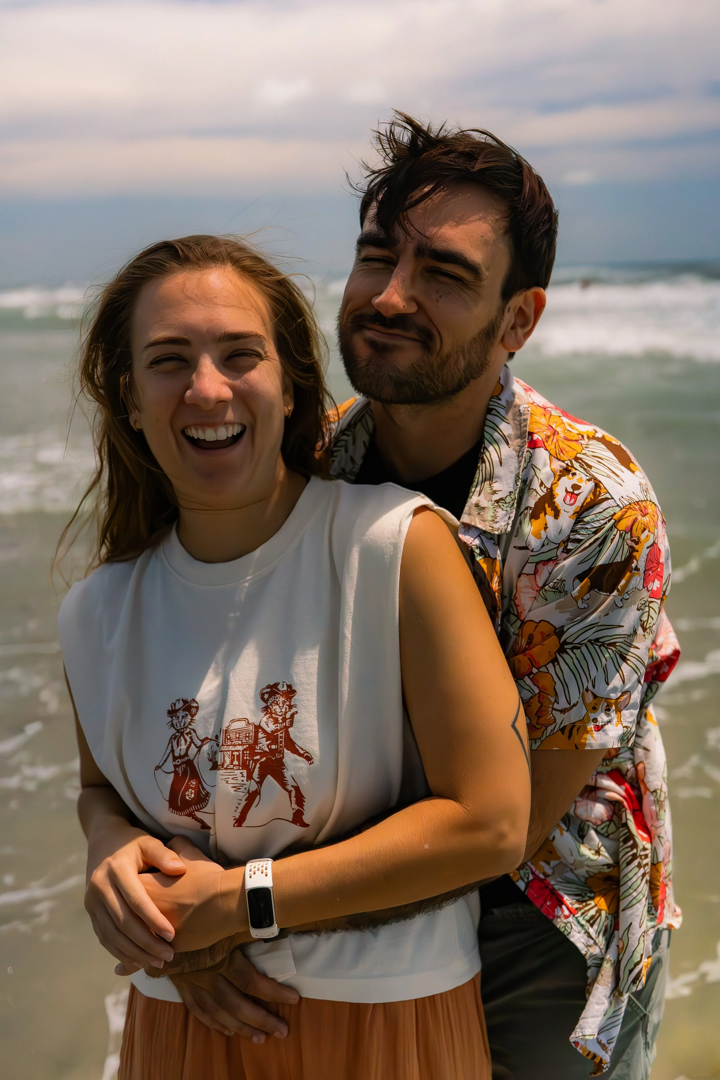 A smiling woman with long hair and a man with a beard and closed eyes at the beach, with waves in the background.