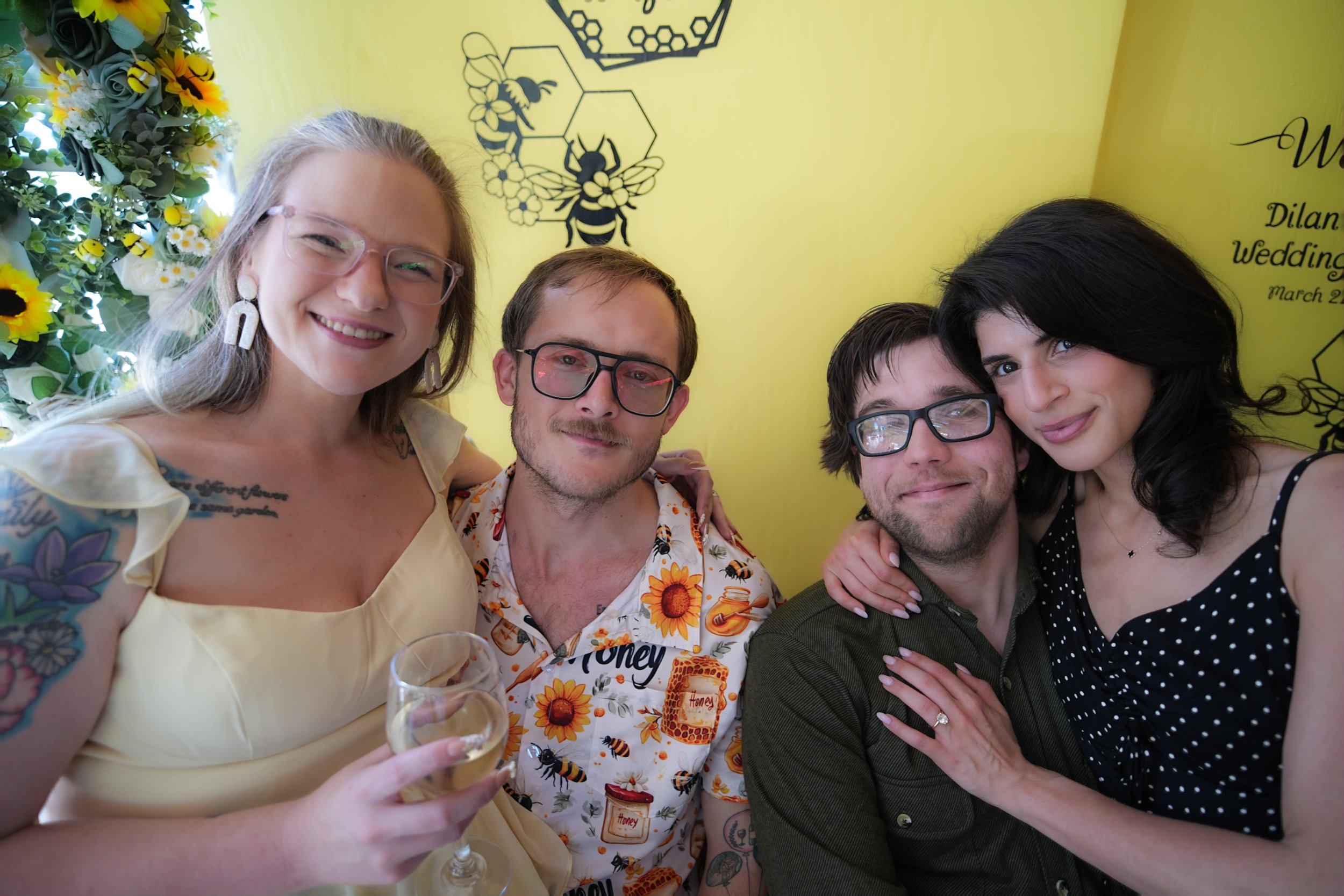 A group of four adults smiling and posing for a photo at a celebration or party, with a yellow wall background decorated with bee and honeycomb illustrations.