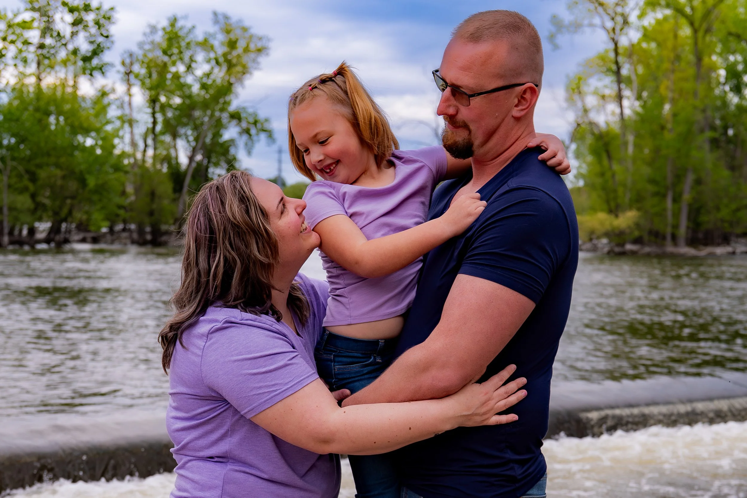 A family of three, a father, mother, and daughter, standing outdoors near a body of water with trees in the background. The daughter is being held by the father and is smiling while looking at her mother, who is holding her around the waist. All are 