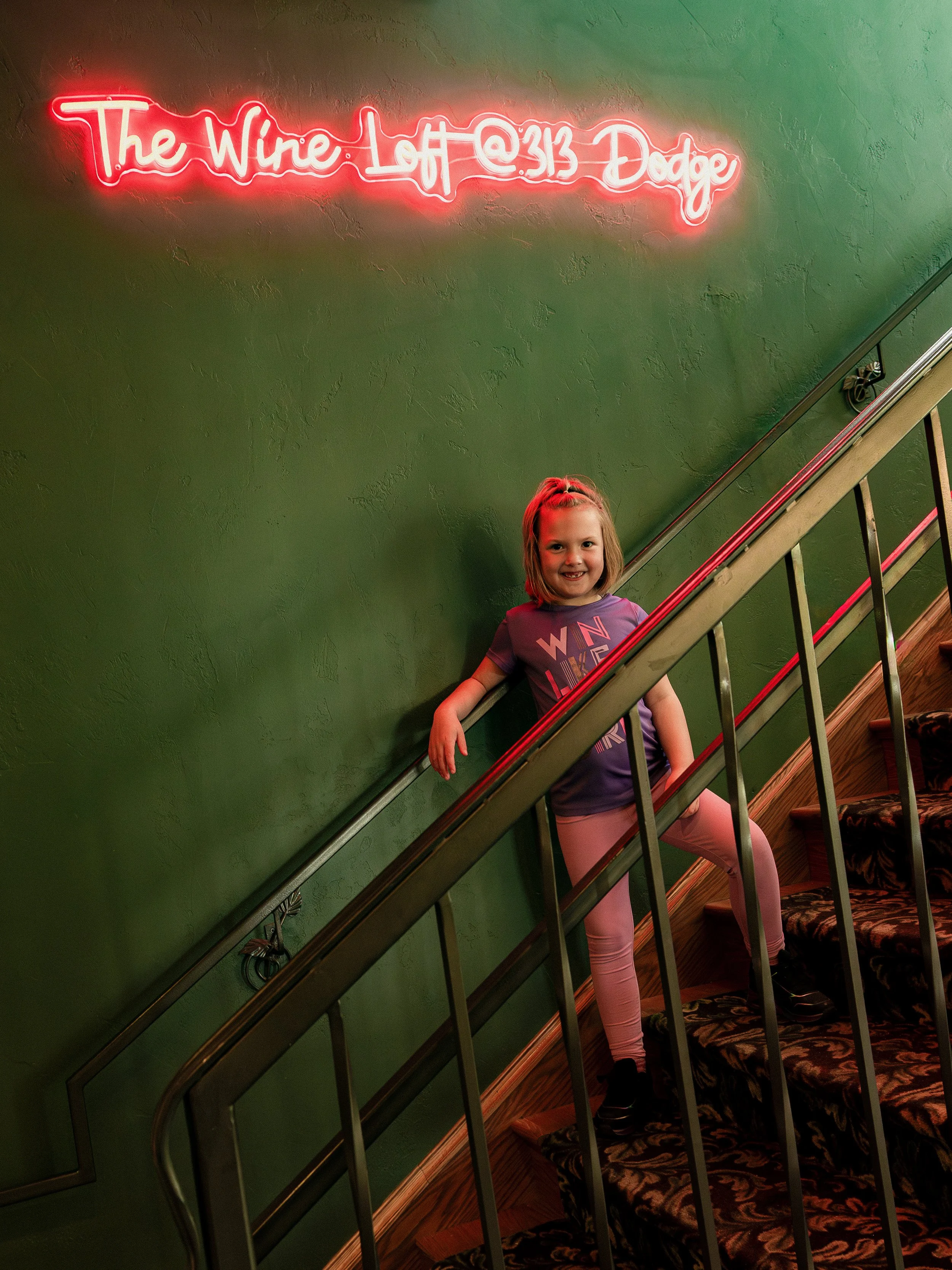 A young girl with blonde hair standing on stairs, smiling, in front of a green wall with a neon sign that reads 'The Wine Loft @313 Dodge'.
