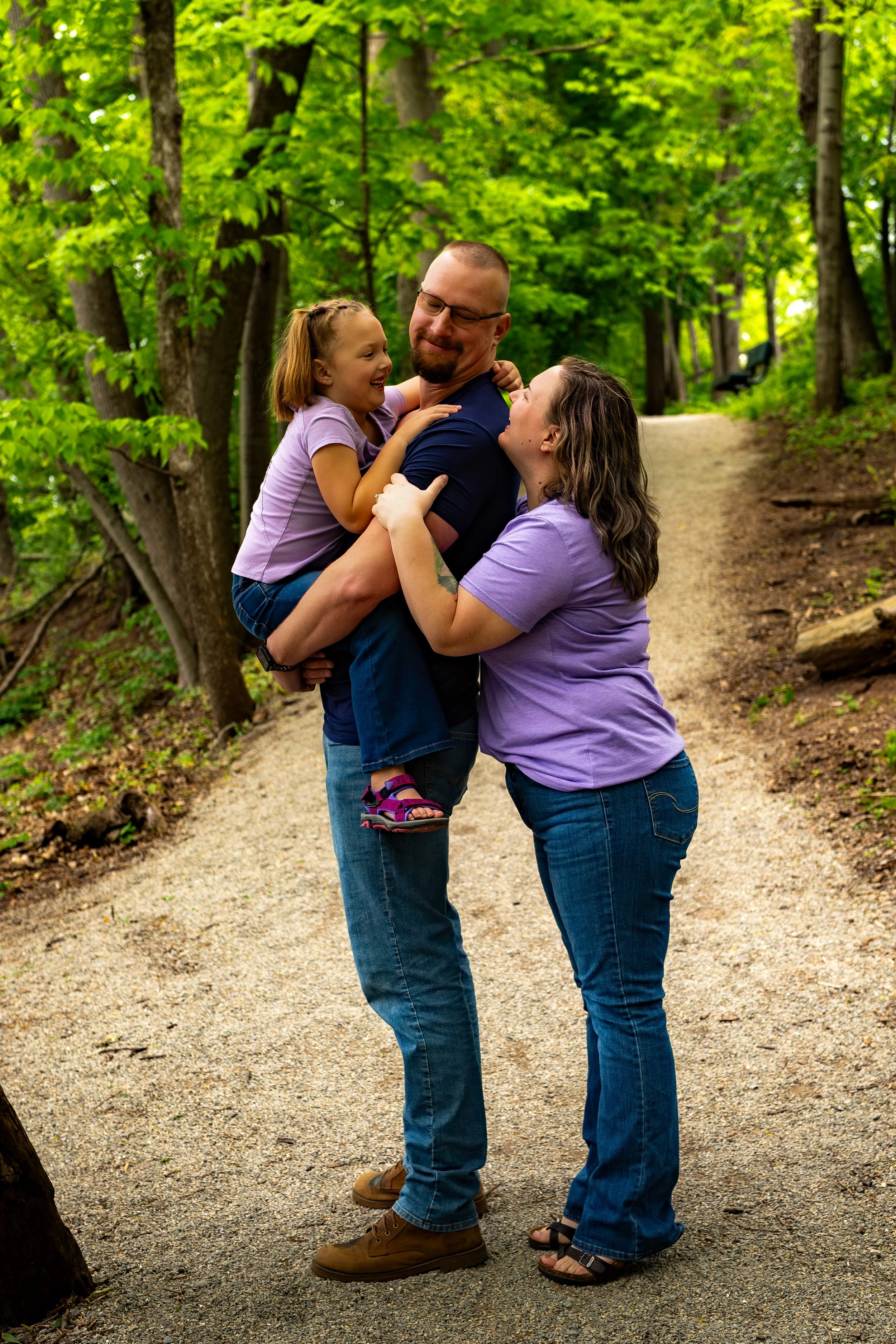 A family of three enjoying a moment outdoors in a wooded park. The man is holding a young girl who is smiling and wearing a purple shirt and shorts. The woman is standing close, smiling up at the girl and man, wearing a purple shirt and jeans. The ba