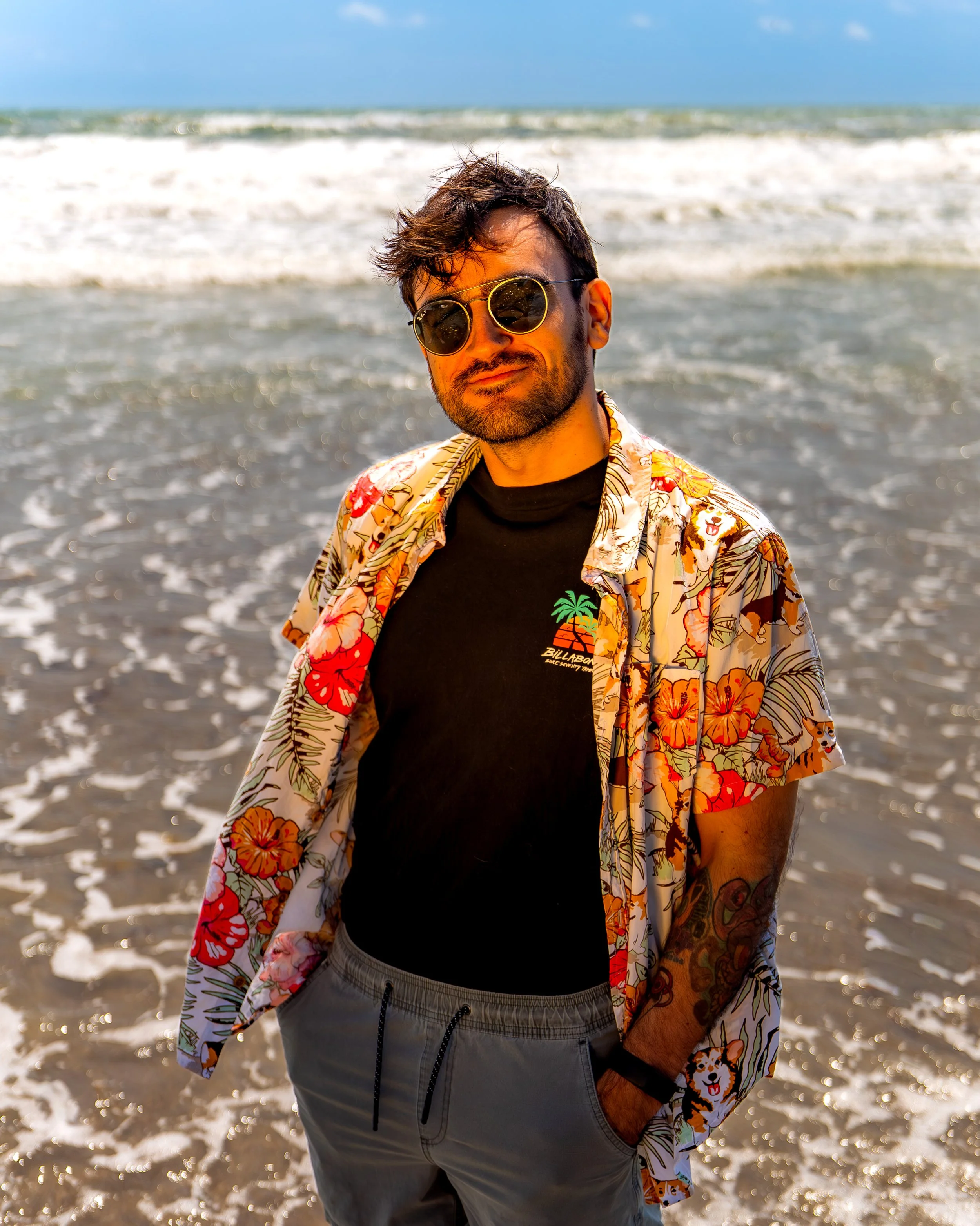 A man with dark hair and a beard wearing sunglasses, a black t-shirt, and a colorful floral shirt, standing on a beach with ocean waves in the background.