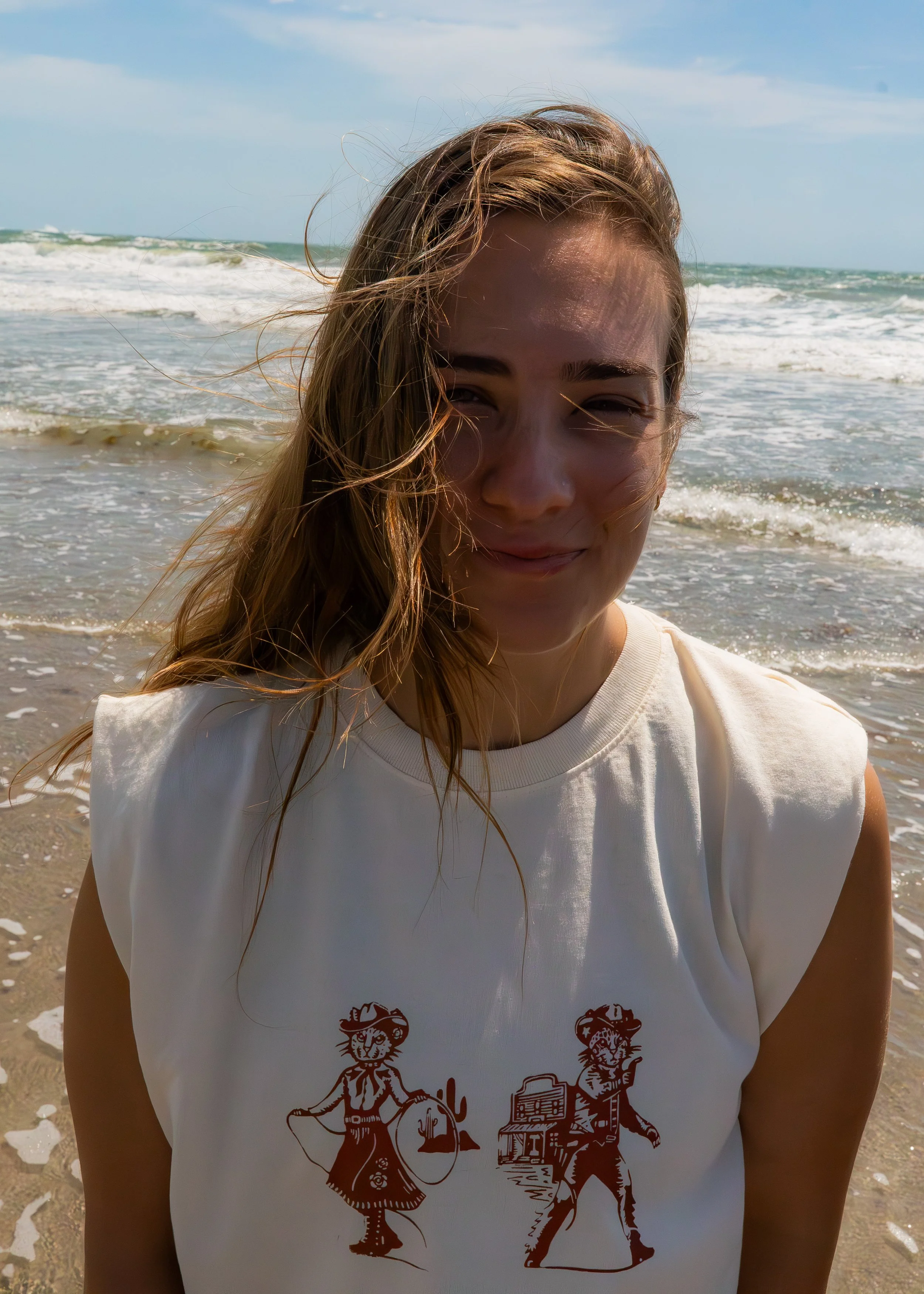 A young girl with long, wet hair, standing on the beach near the ocean, smiling with her eyes closed, wearing a white T-shirt with graphics of lions and a lioness.