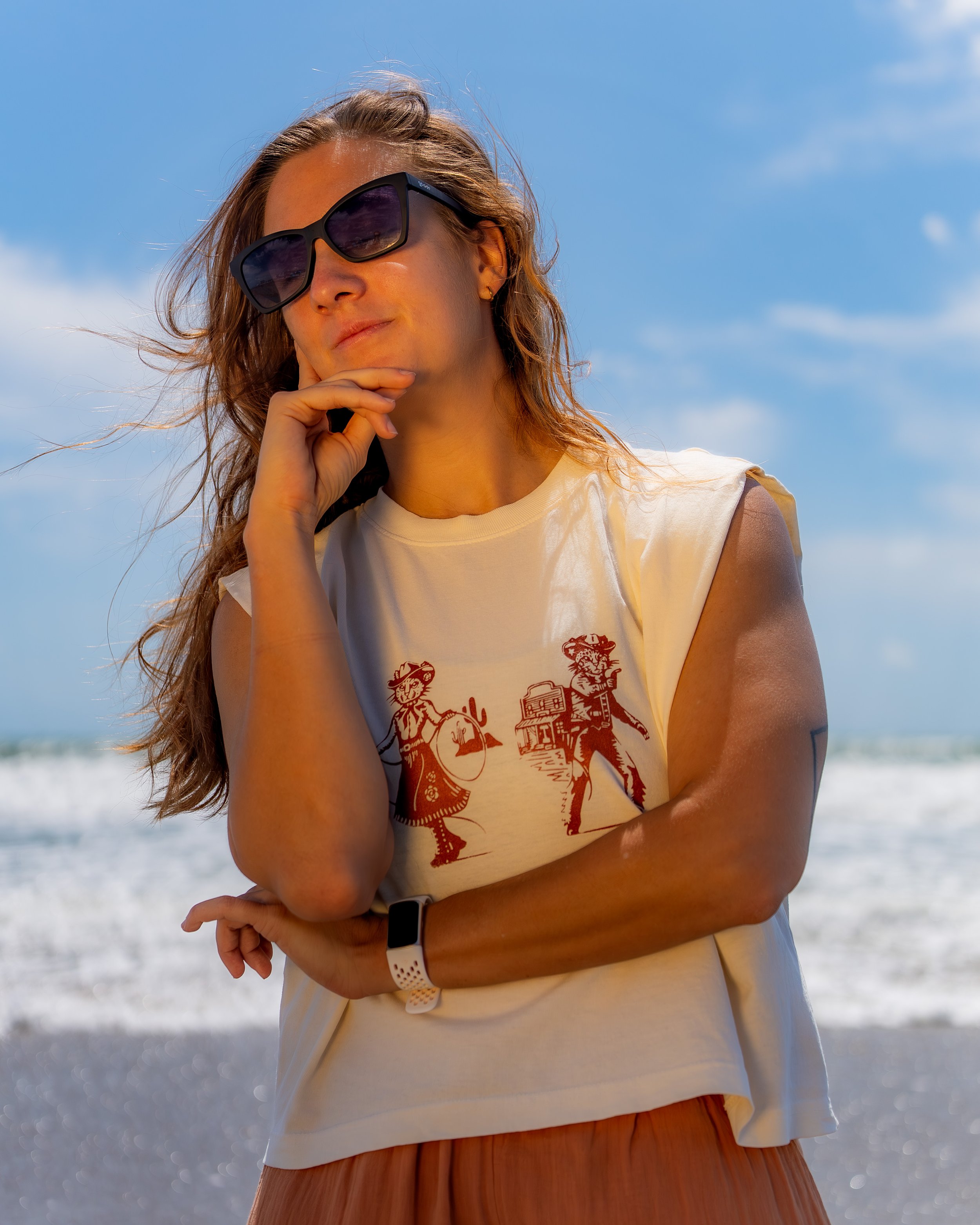 A woman wearing dark sunglasses, a beige sleeveless T-shirt with a graphic print, and a white wristband, standing outdoors near the ocean with her hand resting on her chin, during a sunny day with a blue sky.