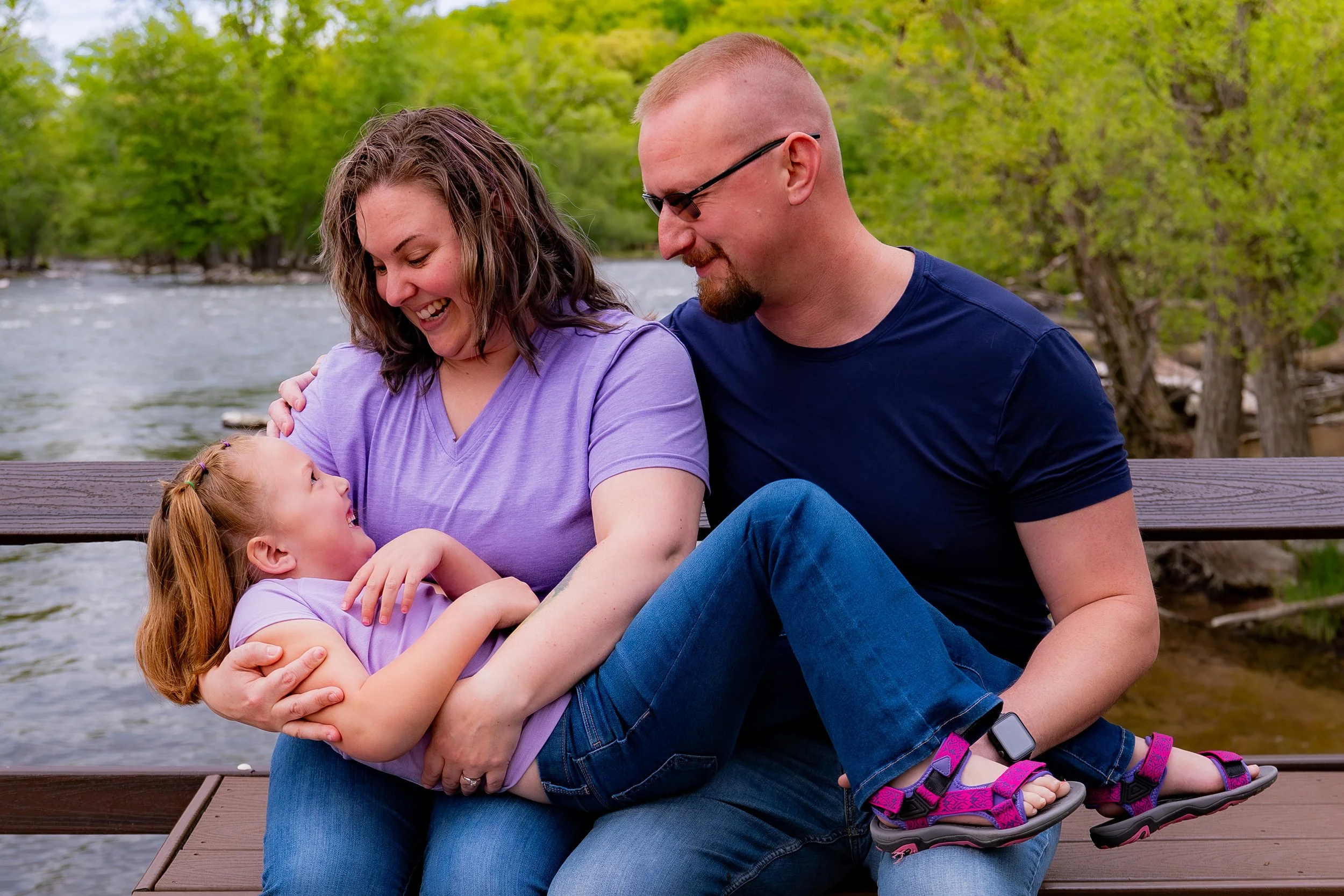 A family of three sitting on a wooden dock by a river in a green, wooded area, smiling and enjoying each other's company.