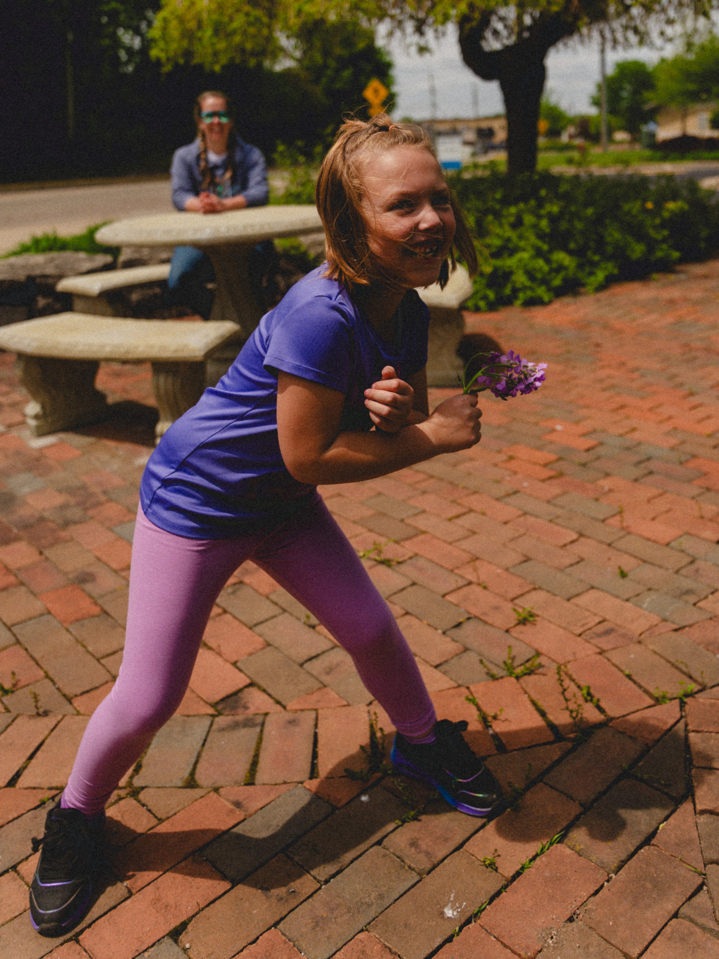 A young girl with light brown hair in a ponytail, wearing a purple shirt and pink leggings, is running outdoors at night while holding a small bouquet of purple flowers. Another girl with glasses and a blue shirt is sitting at a stone table in the ba