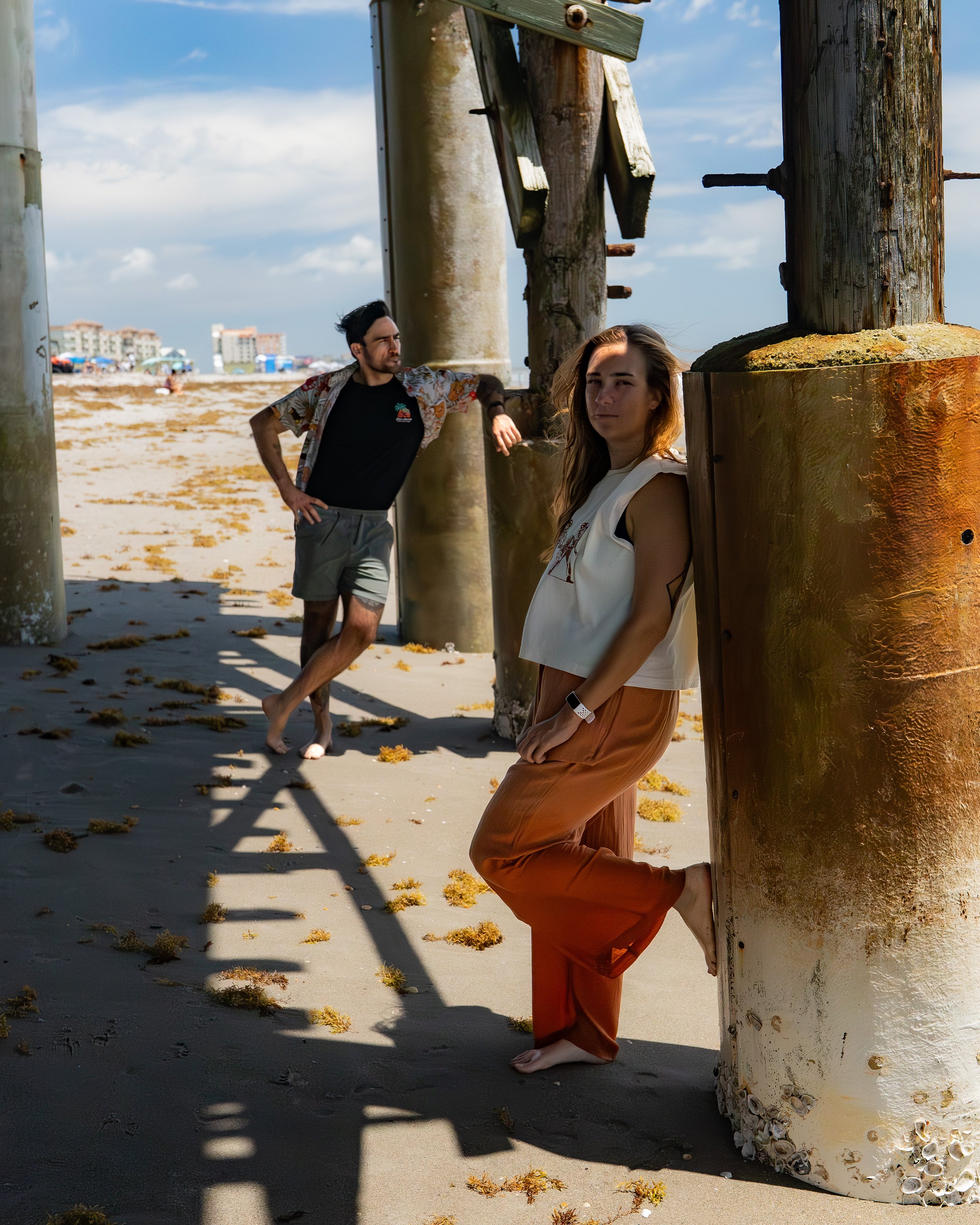 Two young adults, a man and a woman, near the beach with large wooden pilings. The woman is leaning against a pilon with seashells at its base, wearing a white sleeveless top and orange pants, looking at the camera. The man is standing behind another
