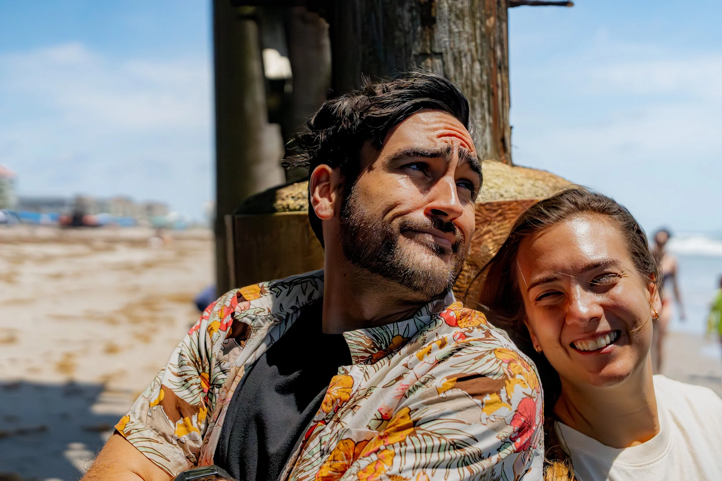 A couple of people, a man and a woman, smiling and standing close together at the beach with a wooden pole behind them and a blue sky overhead. The man has dark hair and a beard, wearing a floral shirt, and partially visible, the woman has light brow