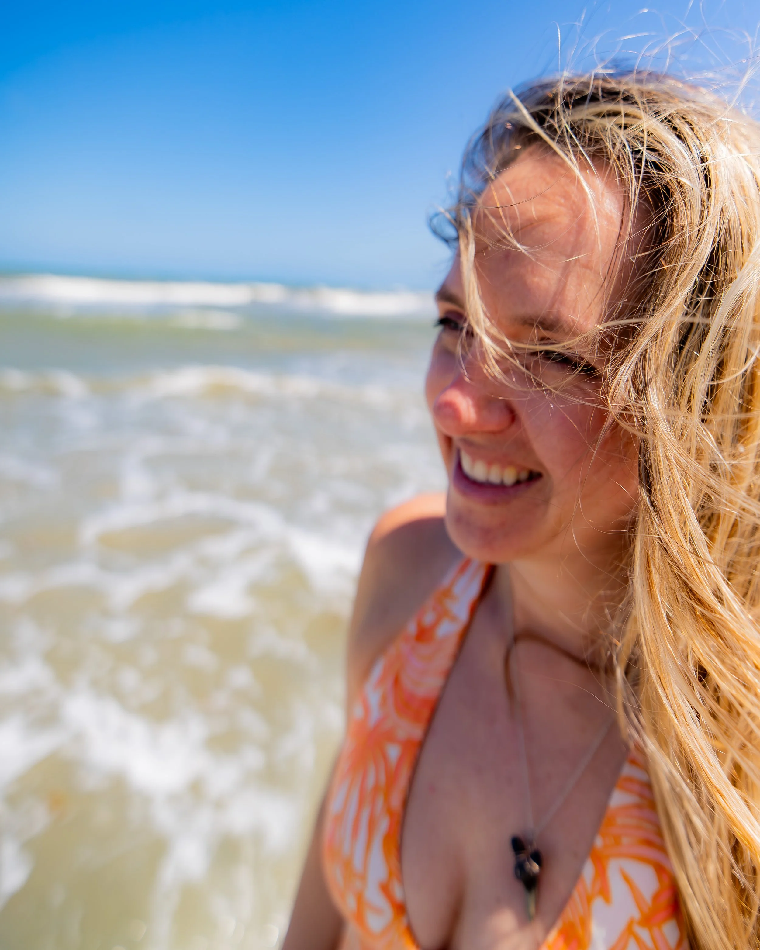 A woman with blonde hair smiling at the beach, wearing an orange and white striped top.