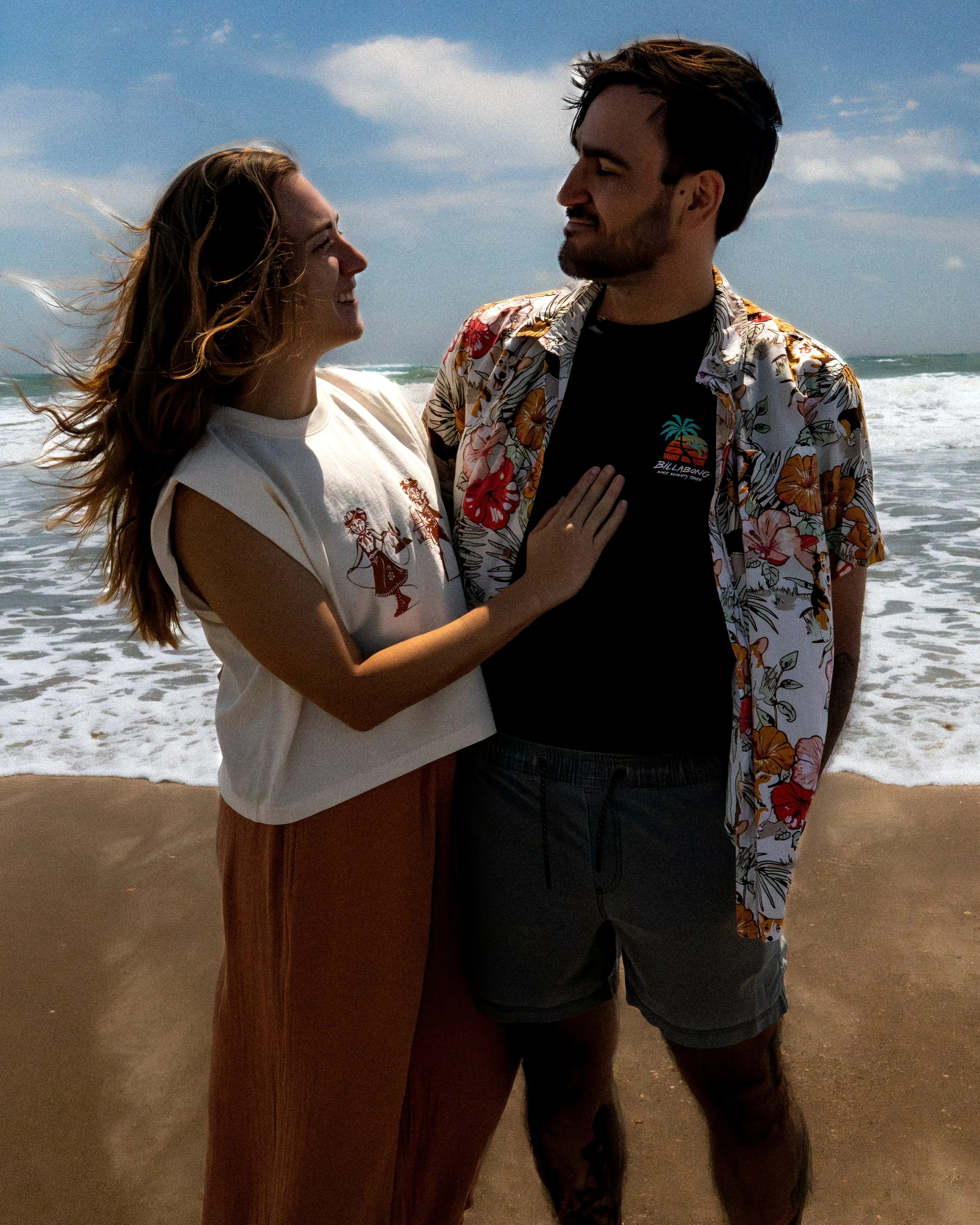 A man and woman standing close together on a beach, facing each other with the ocean and sky in the background. The woman has long, wavy red hair and is smiling at the man. The man has short dark hair, a beard, and is smiling back at her. The woman i