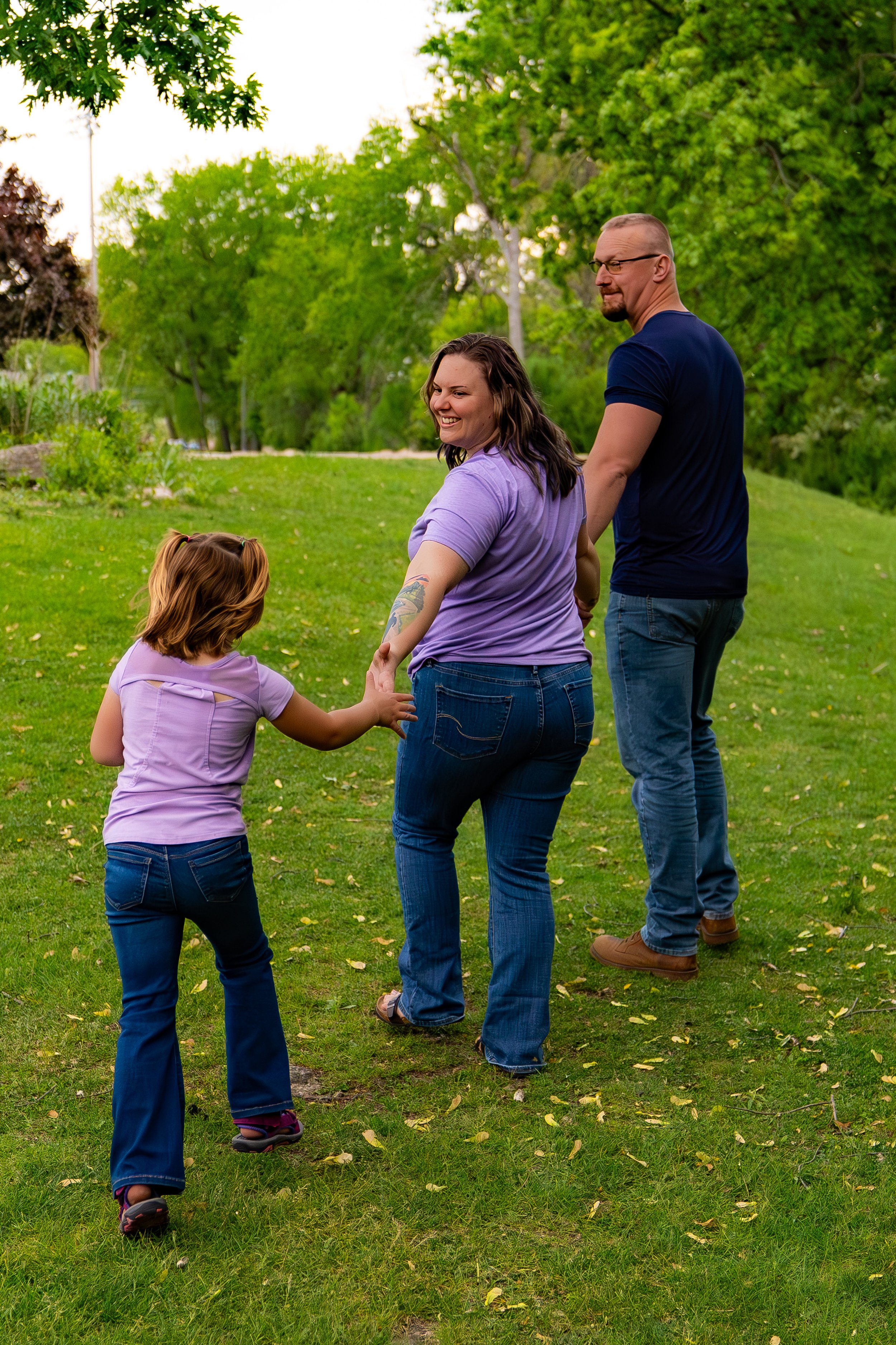 A family of four, including a young girl, a woman, and a man, holding hands and walking in a park with lush green trees and grass, during daytime.