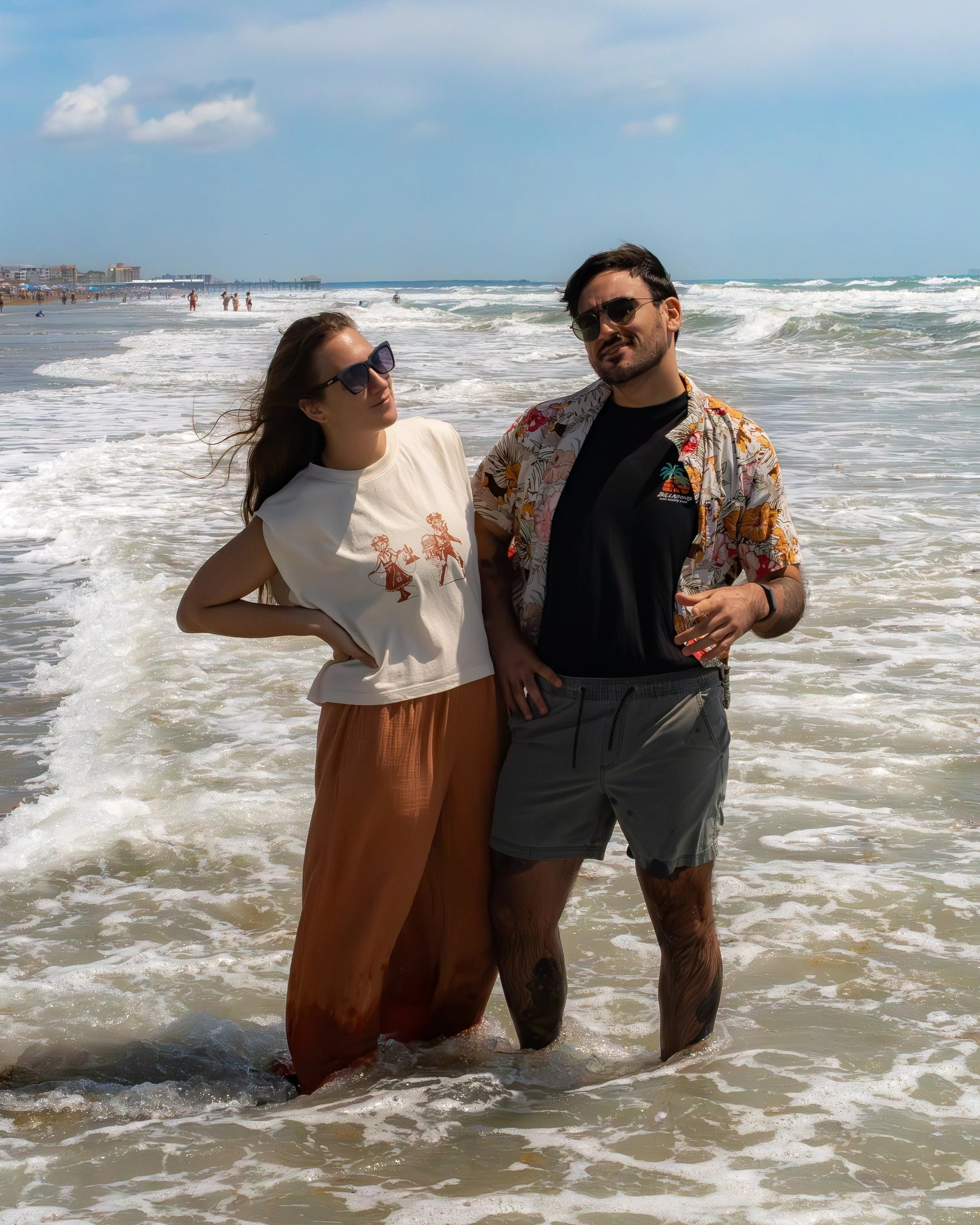 A couple standing in the shallow ocean water at the beach, smiling and wearing sunglasses, with a city pier and beachgoers in the background on a sunny day.