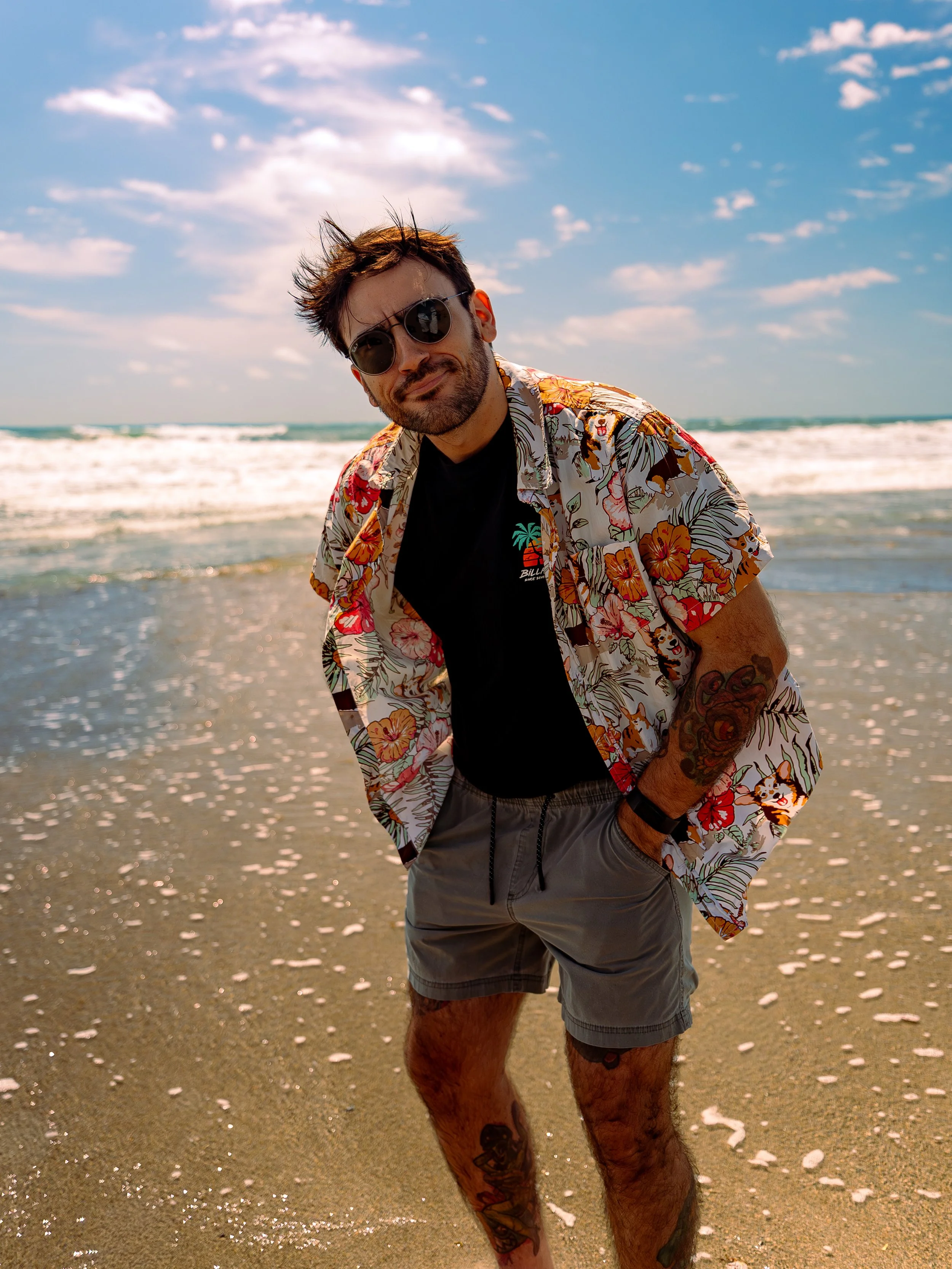 A man with dark hair and a beard wearing sunglasses, a floral Hawaiian shirt over a black T-shirt, and gray shorts, standing on the beach with waves and a partly cloudy sky in the background.