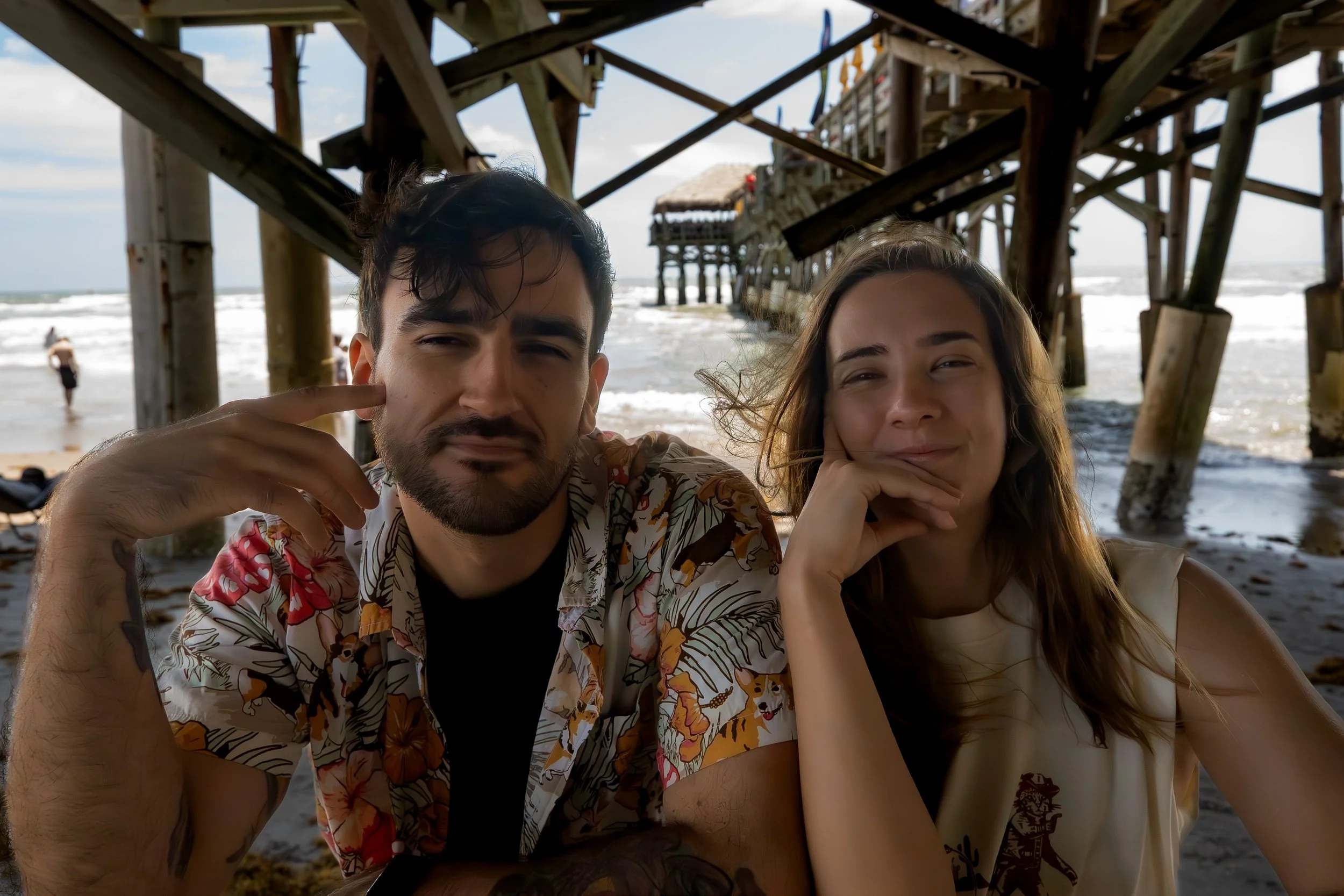 Two young adults, a man and a woman, posing together under a pier at the beach. The man has dark hair, facial hair, and is wearing a tropical shirt. The woman has long light brown hair and is resting her chin on her hand. The pier's wooden structure 