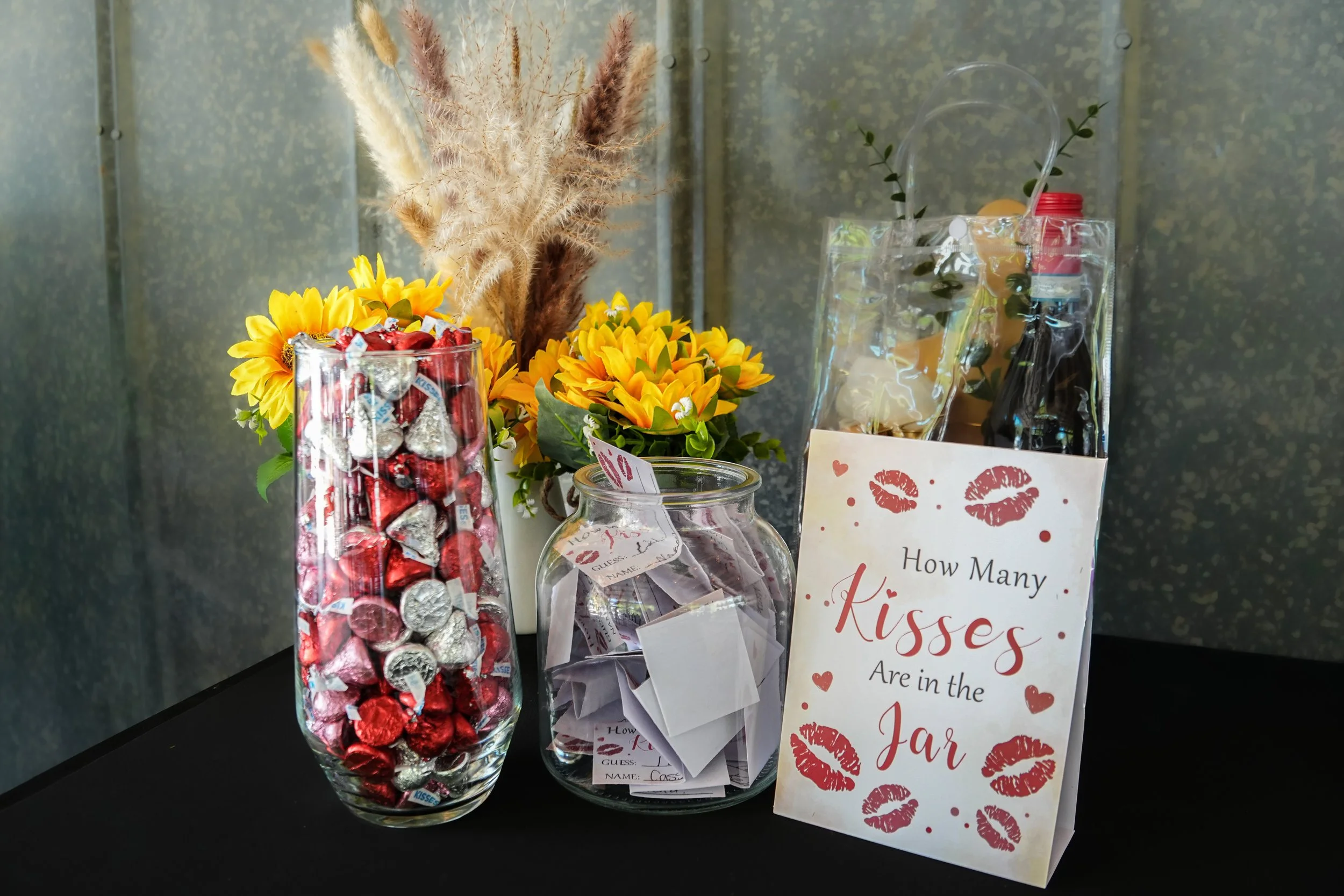 A tall glass jar filled with foil-wrapped candies next to yellow flower arrangements and a small sign that reads "How Many Kisses Are in the Jar" with red kiss marks and a red lip print.