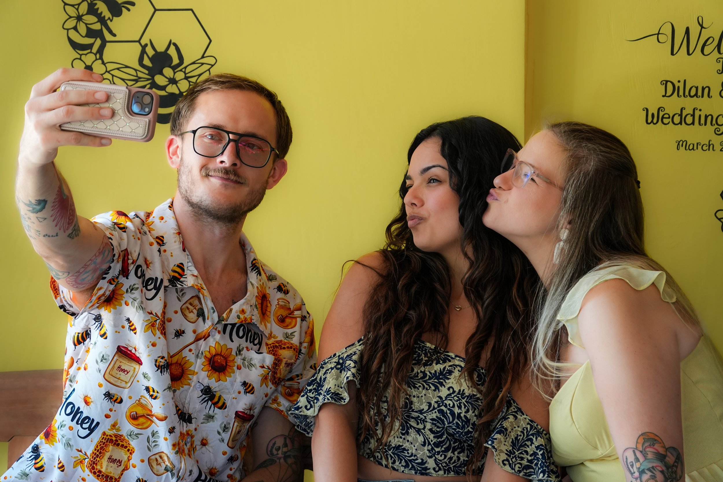Three women and one man taking a selfie together against a yellow wall with a bee and honeycomb decoration. The man is holding the phone, and all are smiling or making playful faces.