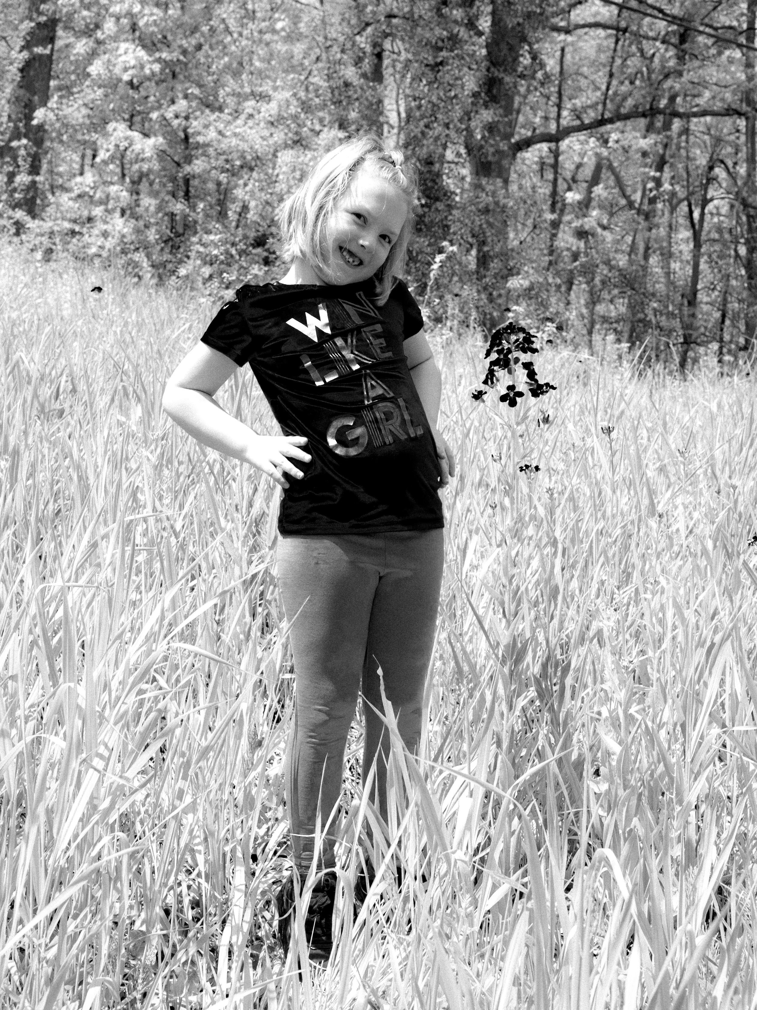 A young girl smiling and posing in a grassy field with trees in the background.