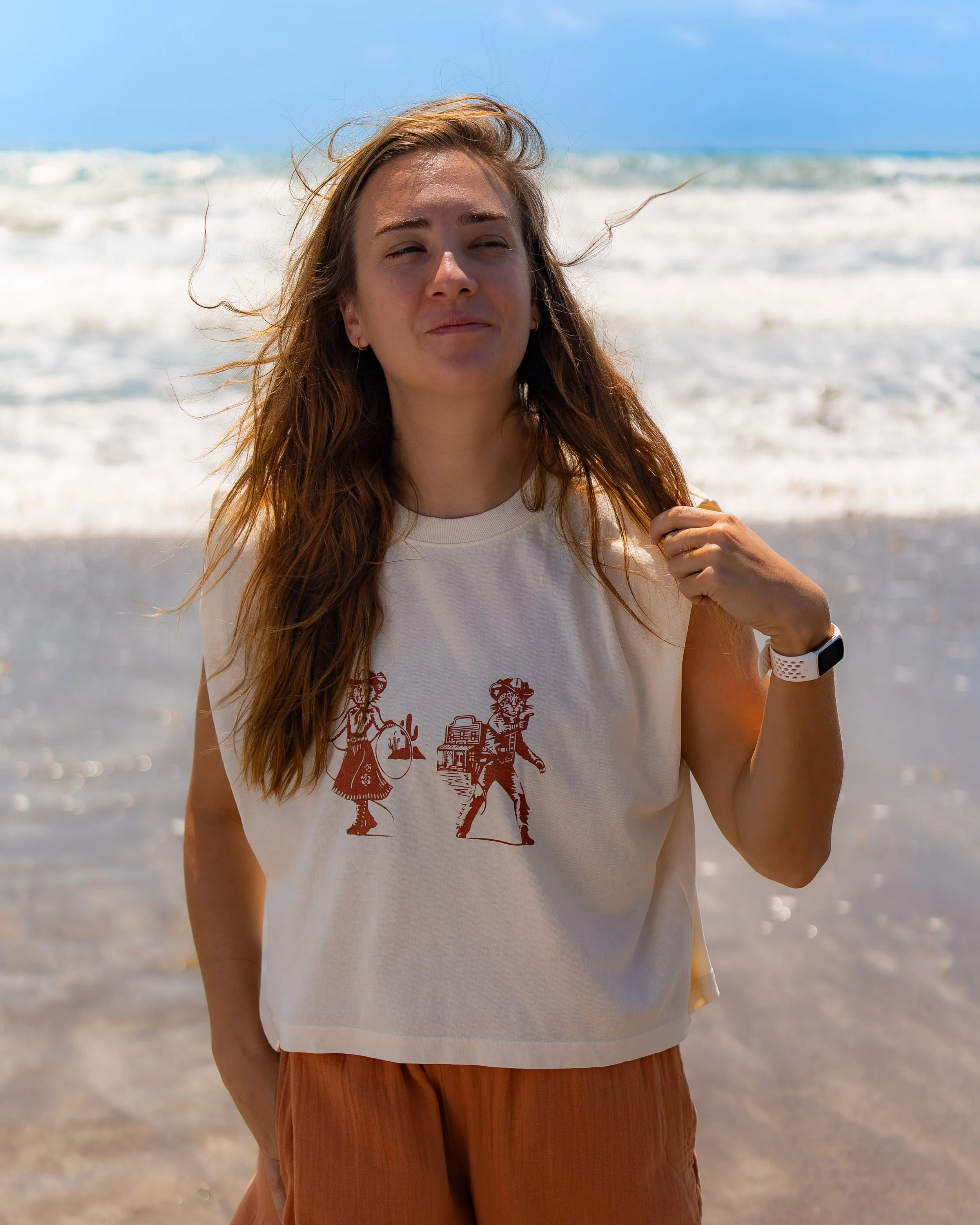 A woman with long, light brown hair stands on a beach with the ocean in the background. She is wearing a white t-shirt with a red graphic and brown pants, holding her hair with one hand, and has a watch on her wrist. The sky is partly cloudy, and wav