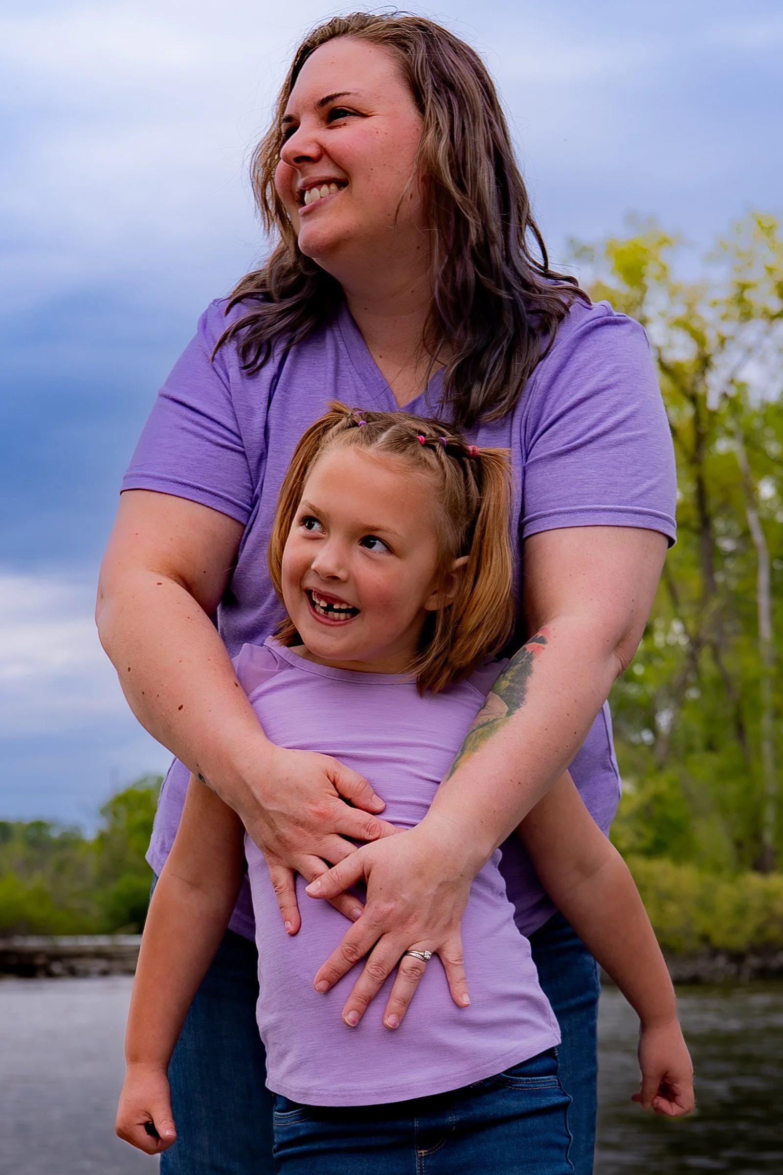 A woman and a girl are outdoors, smiling and hugging. The woman has shoulder-length brown hair, is wearing a purple shirt, and has a tattoo on her left arm. The girl has red hair with two pigtails, is also wearing a purple shirt, and is smiling with 