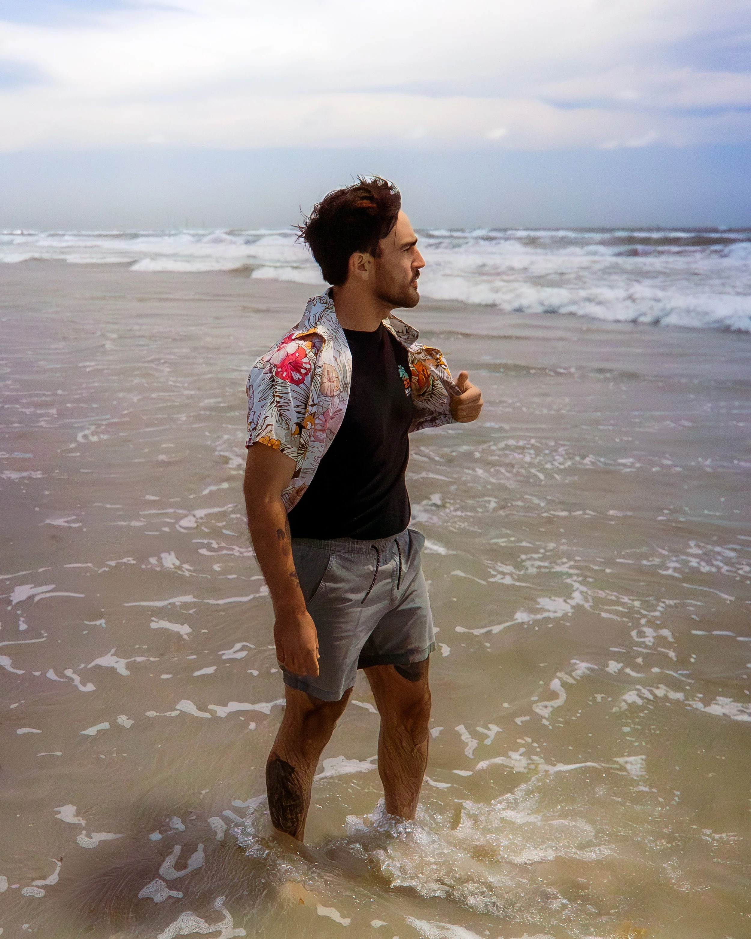 A man with dark hair and a beard standing knee-deep in ocean water at the beach, wearing a floral shirt over a black t-shirt, looking to the right, with waves and cloudy sky in the background.