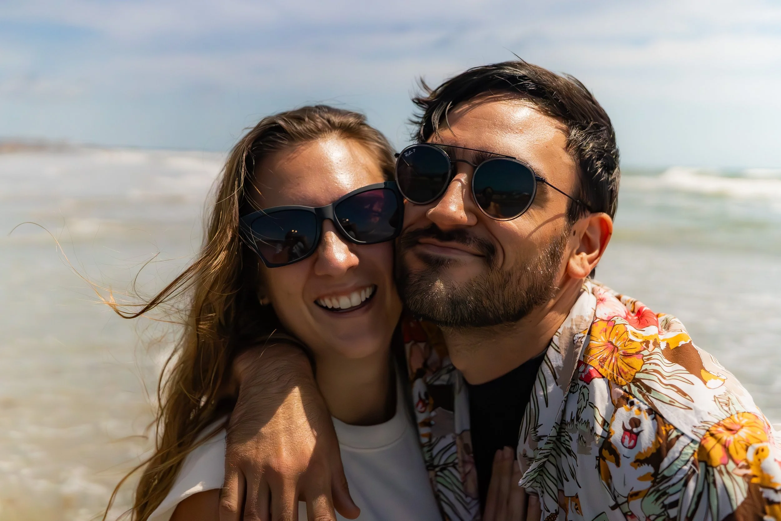 A smiling couple wearing sunglasses taking a selfie on the beach with ocean waves in the background.