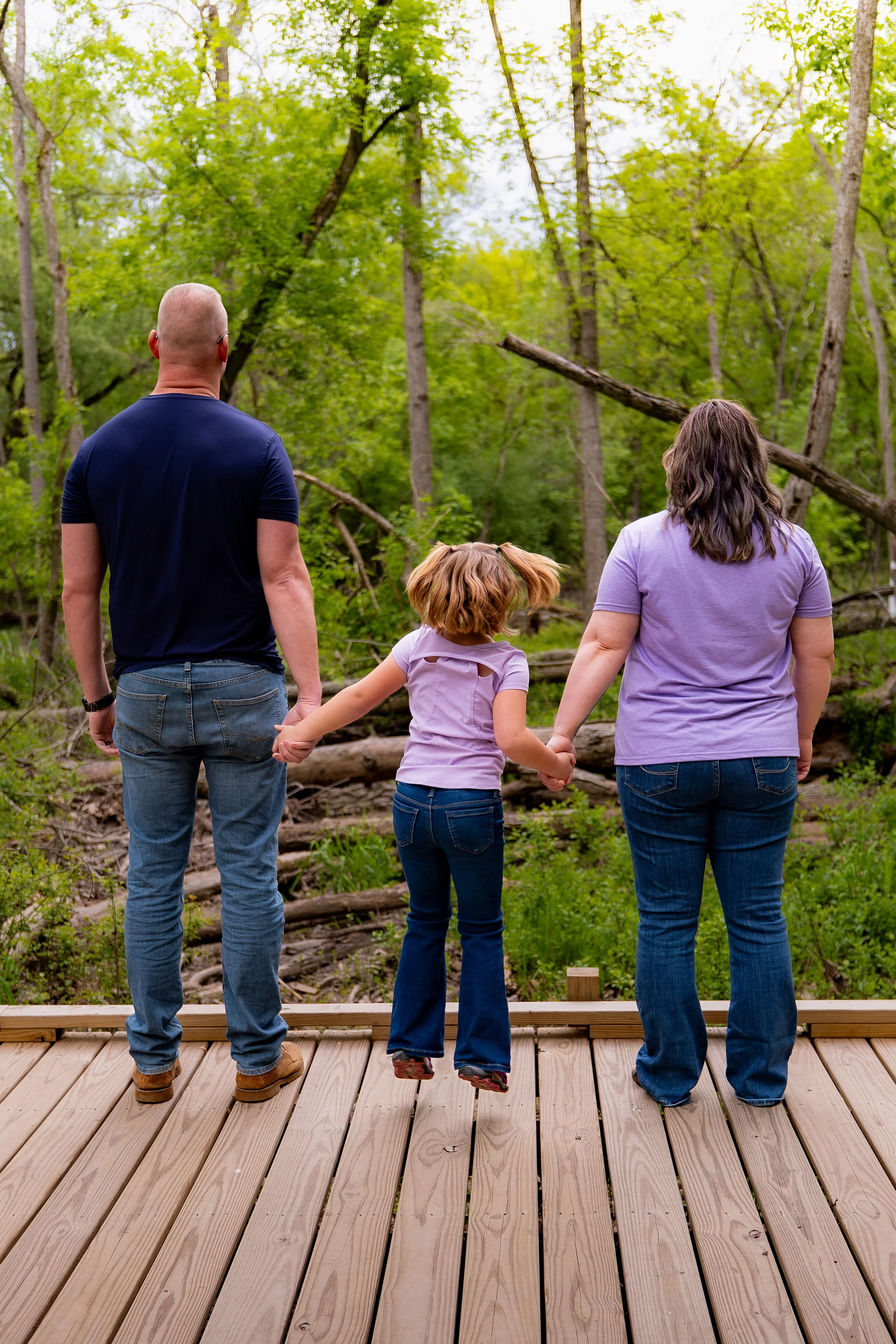A family of three, two adults and a child, holding hands and standing on a wooden deck, looking at a fallen tree in a green forest.