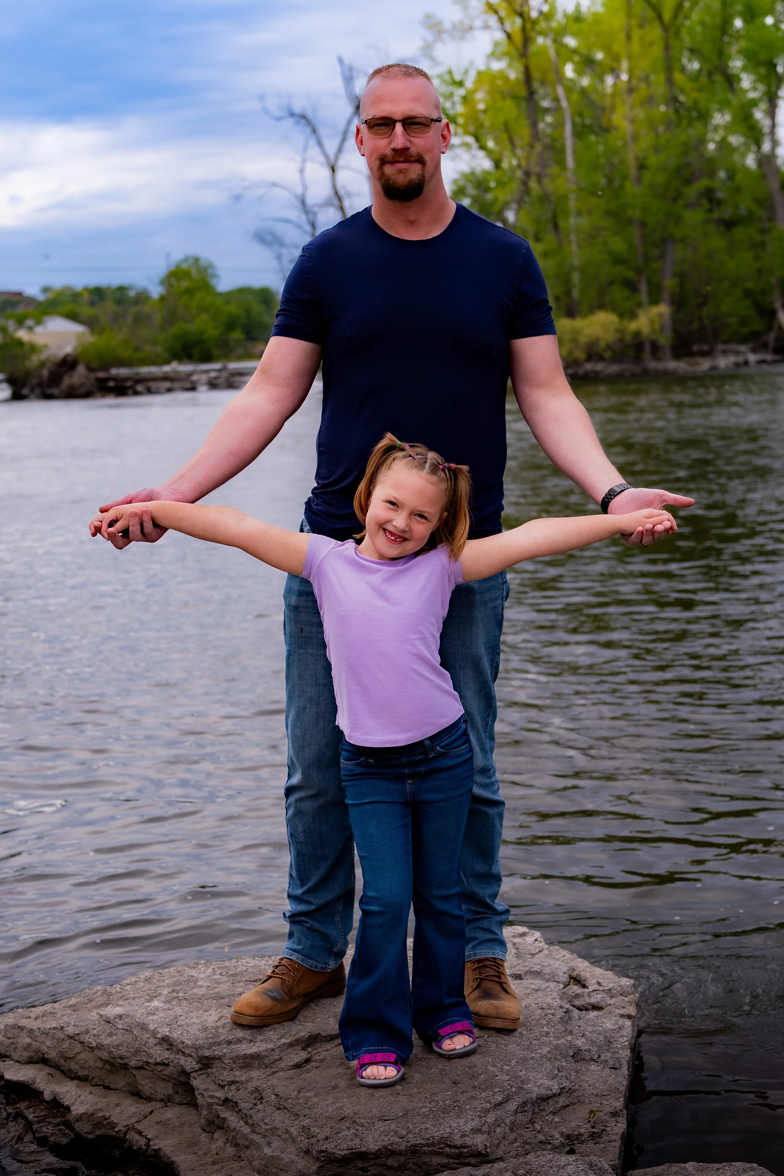 A man and a young girl standing on a rock by a body of water, with trees and a cloudy sky in the background.