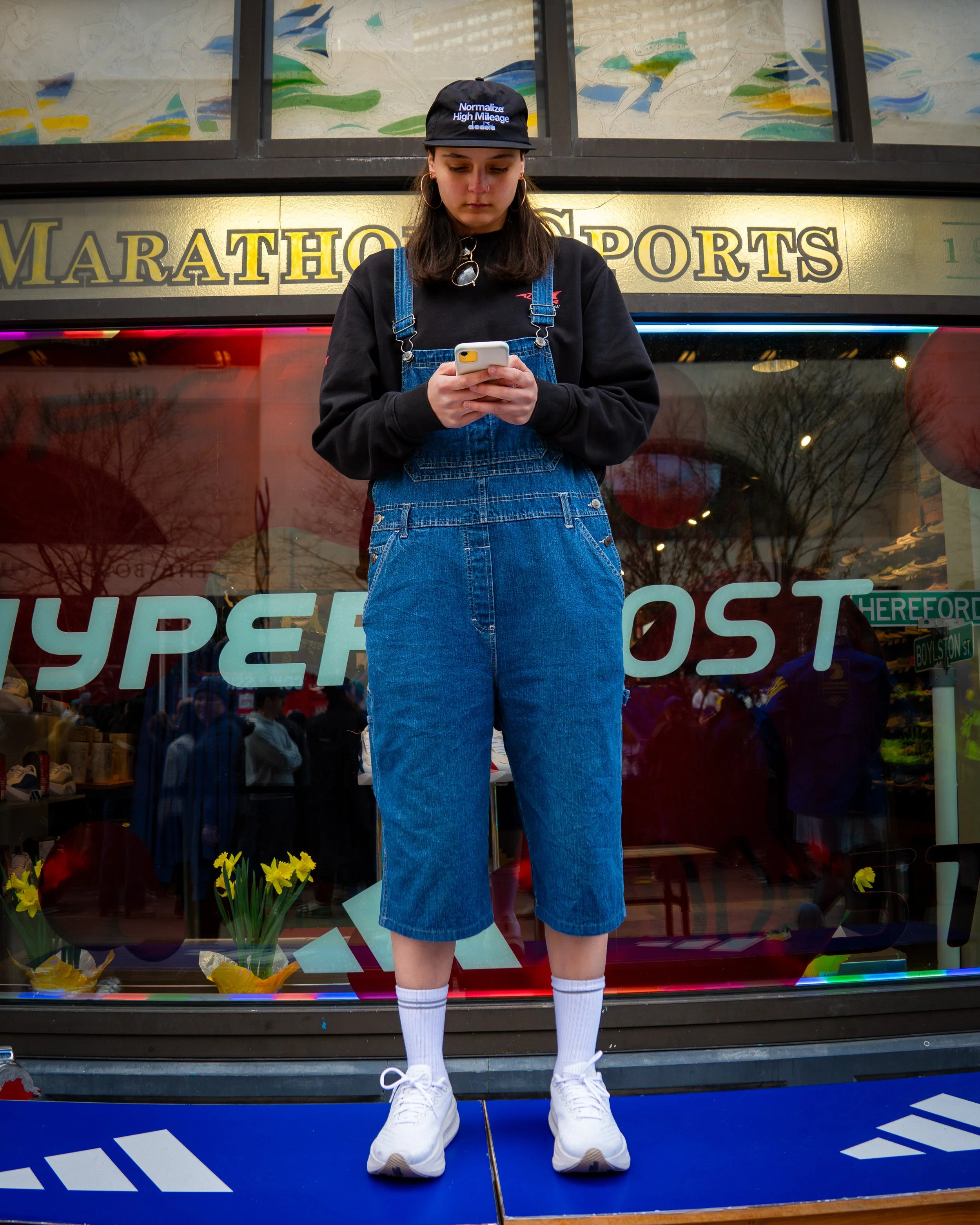 A young woman standing inside a store window, looking at her phone. She is wearing a black cap, black sweatshirt, denim overalls, white socks, and sneakers. The store window behind her displays the words 'MARATHON SPORTS' and other signs, with reflec
