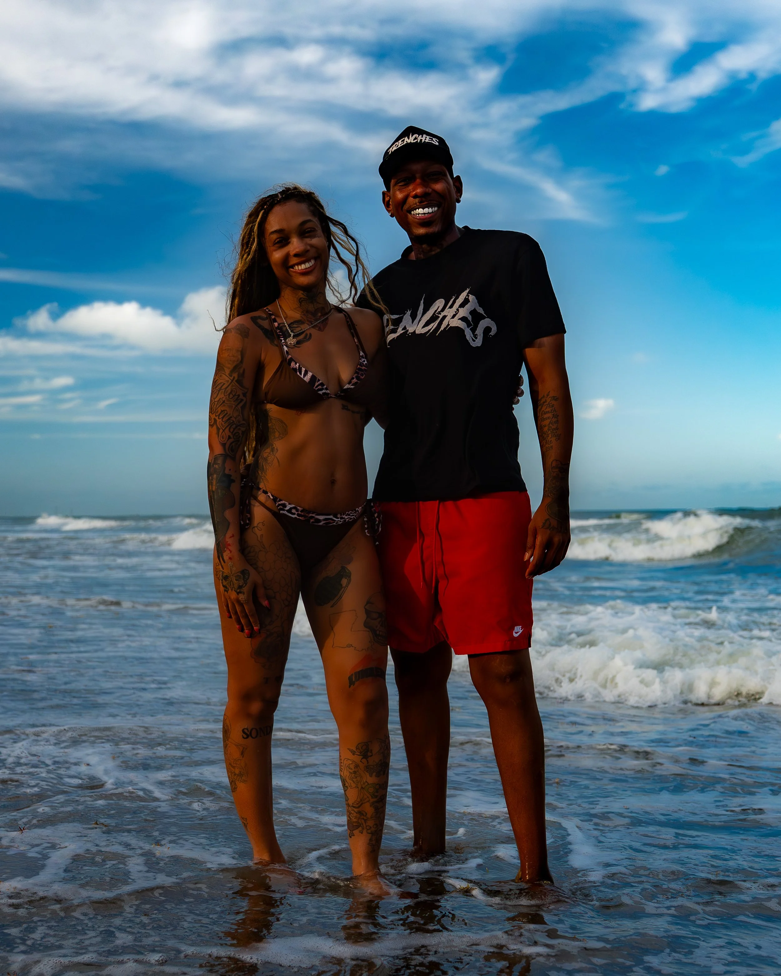 A smiling woman and man standing in shallow ocean water with waves in the background under a partly cloudy sky, both with numerous tattoos and casual beachwear.