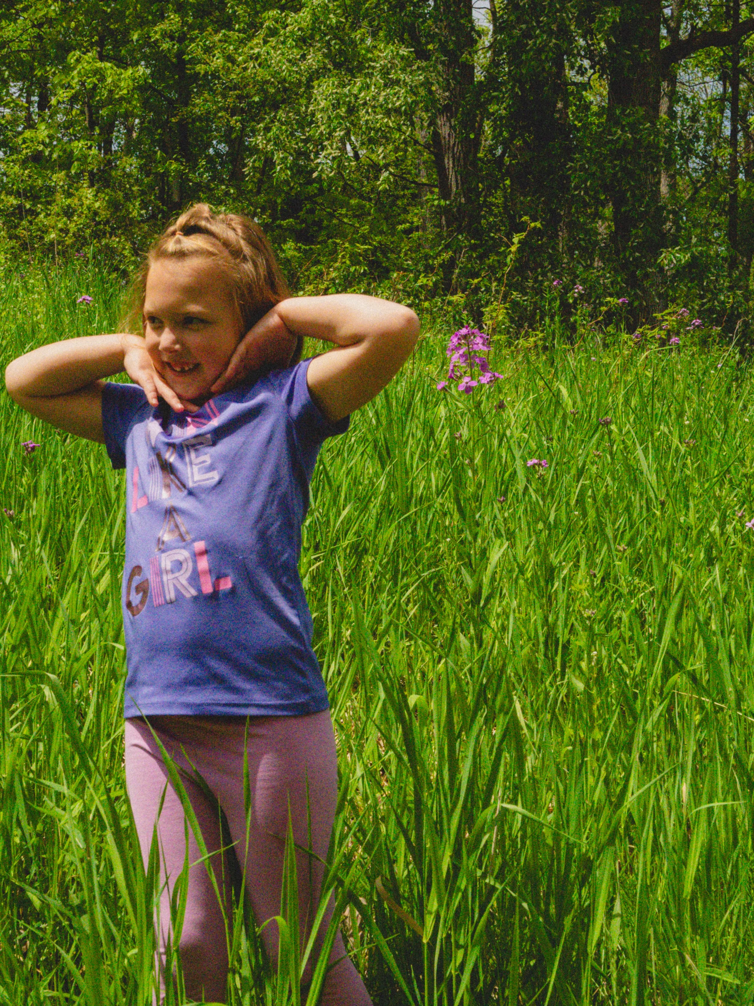 A young girl with blond hair is standing in a grassy field with pink flowers, smiling and posing with her hands near her face, surrounded by lush green trees under bright sunlight.