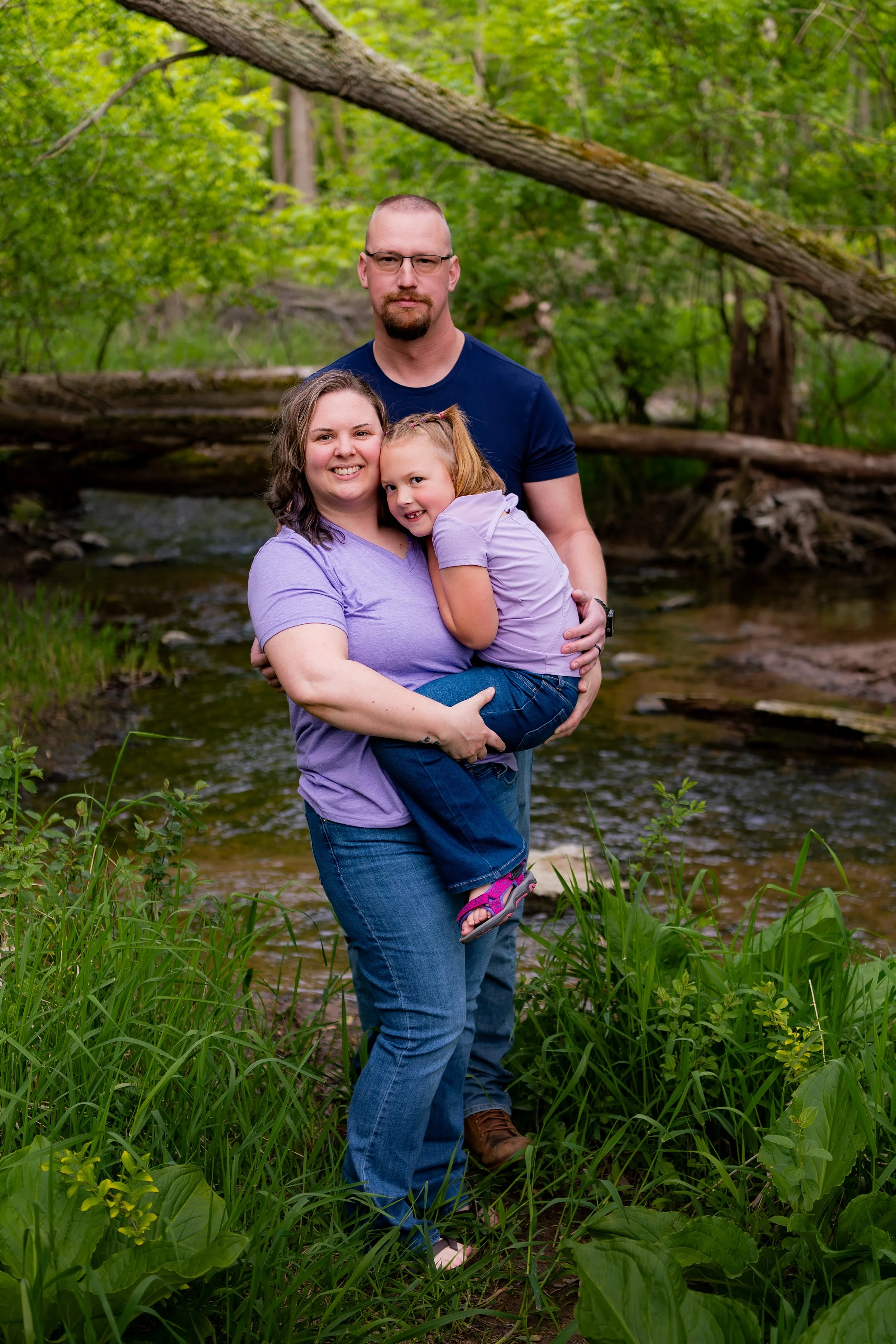 A family of three standing in a lush green forest by a creek, embracing and smiling at the camera. The mother is holding her daughter, and the father stands behind them with a serious expression.