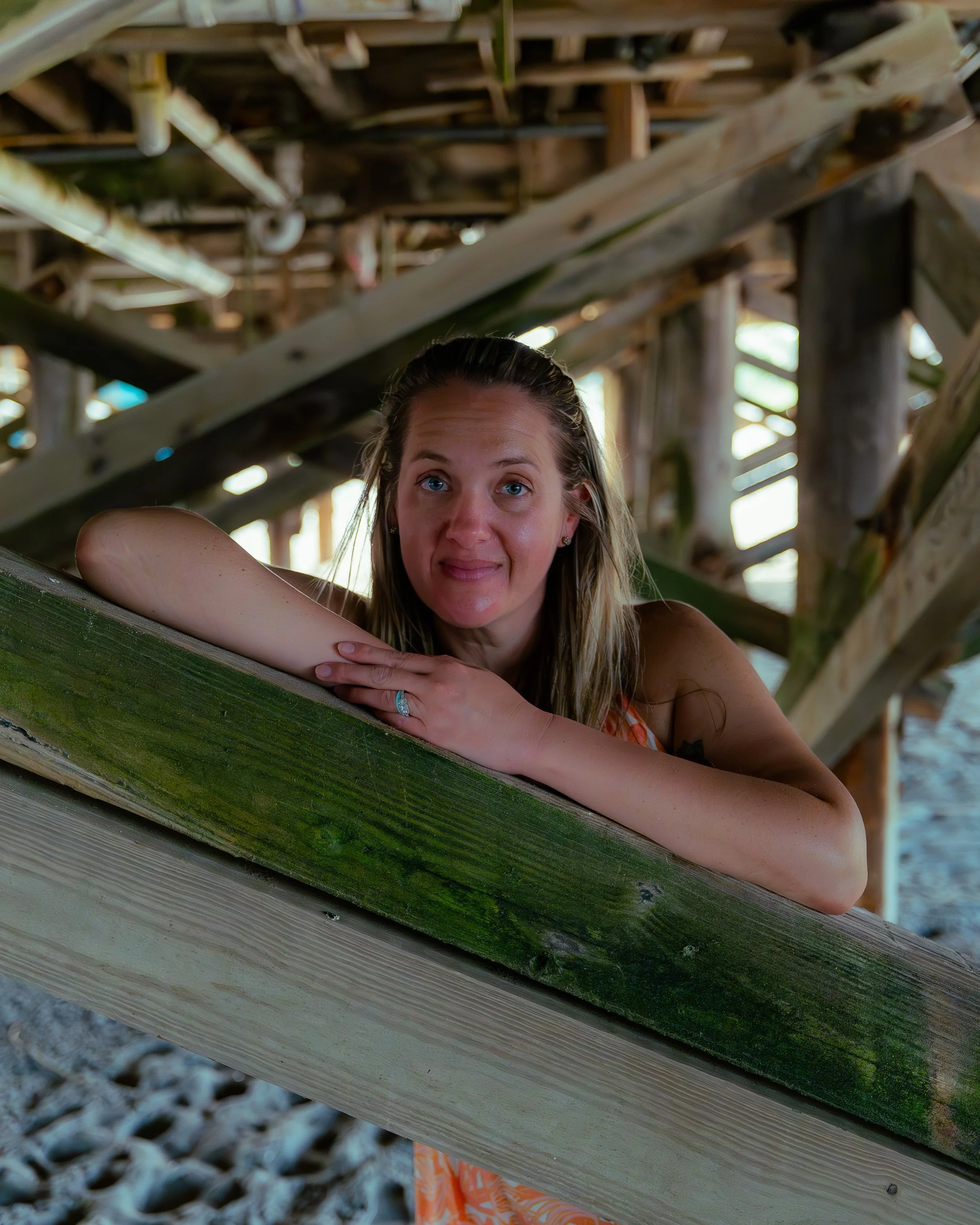 A woman with blue eyes and long blonde hair leaning on a wooden railing outdoors, with a background of wooden structure and greenery.