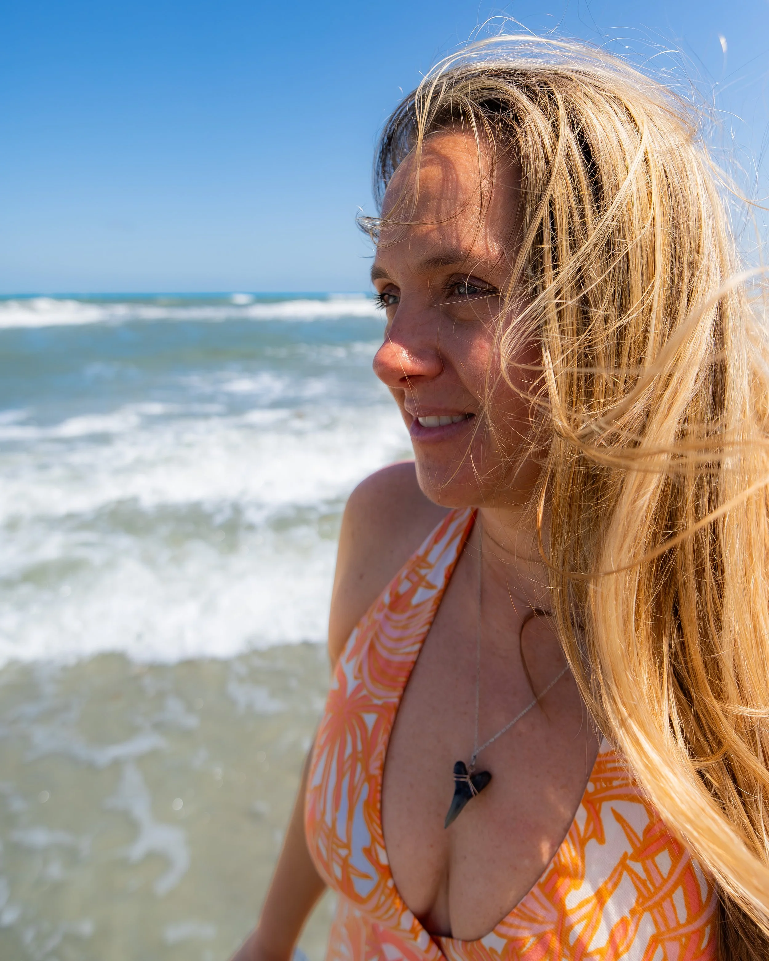 A young woman with long blonde hair smiling by the ocean, wearing an orange and white patterned dress and a black necklace, with waves in the background.