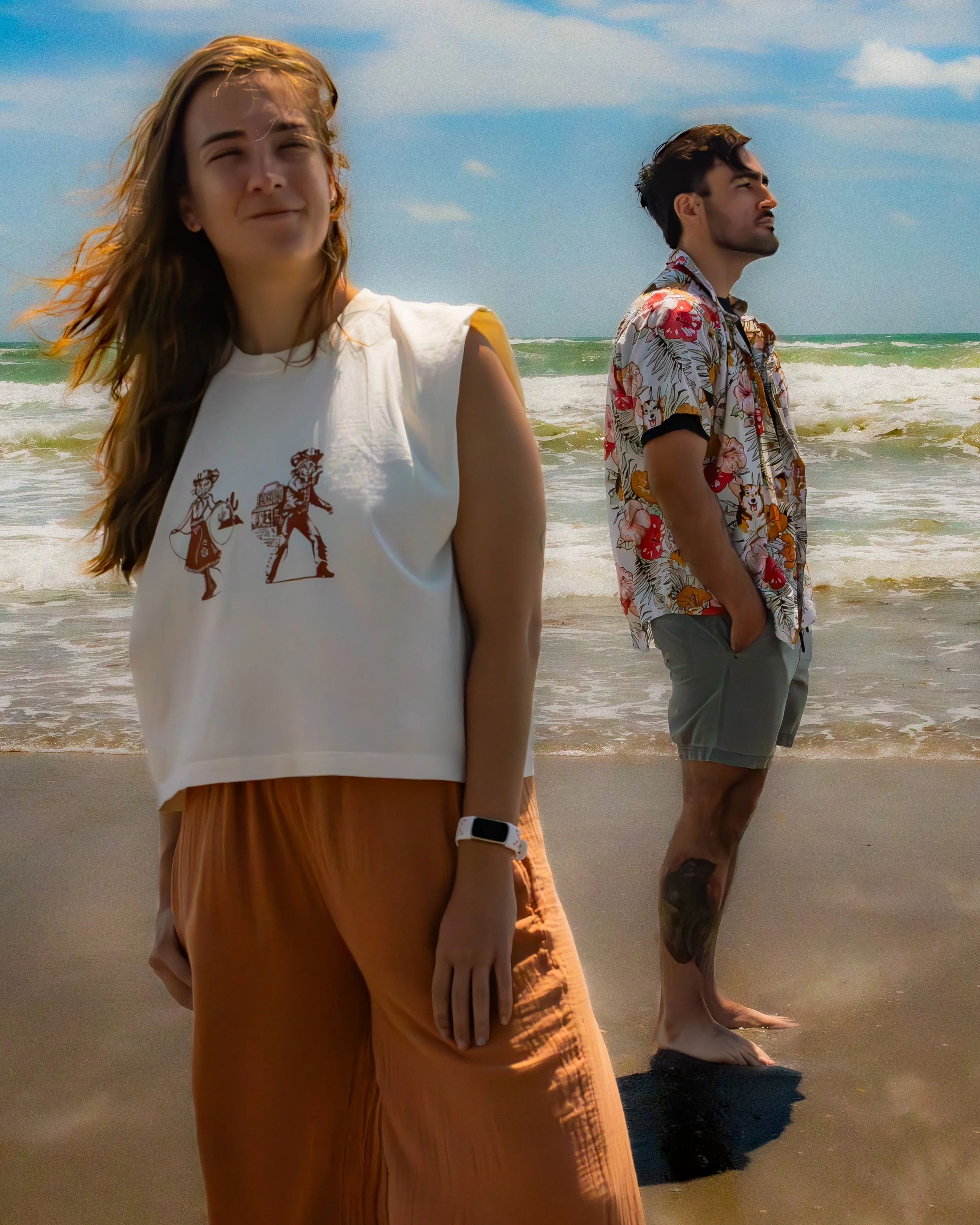 Two people standing on the beach near the water, one woman and one man, both facing to the right. The woman is in the foreground wearing a white sleeveless top with a graphic design and a watch, and the man is in the background wearing a colorful Haw