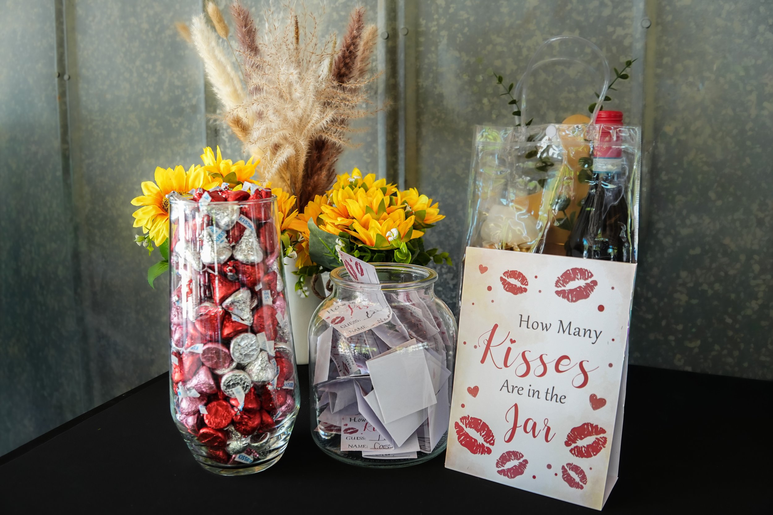 Close-up of a glass jar filled with assorted individually wrapped candies, next to a small bouquet of yellow flowers and a sign with red lipstick kiss marks and the text 'How Many Kisses Are in the Jar'.