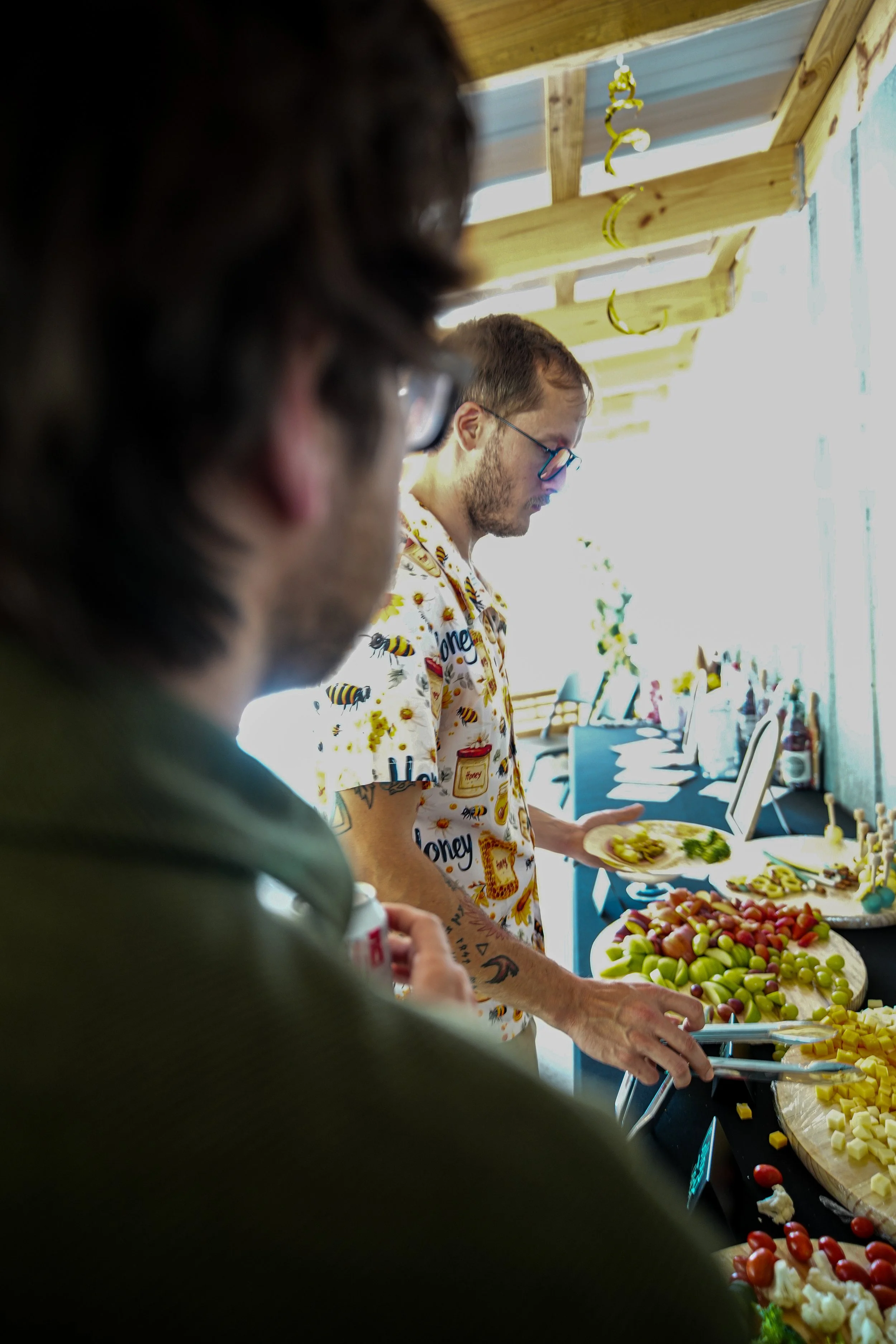 A person with glasses, wearing a shirt with honey and bee patterns, serving food at a buffet table with various vegetables and dishes. In the foreground, another person is partially visible.