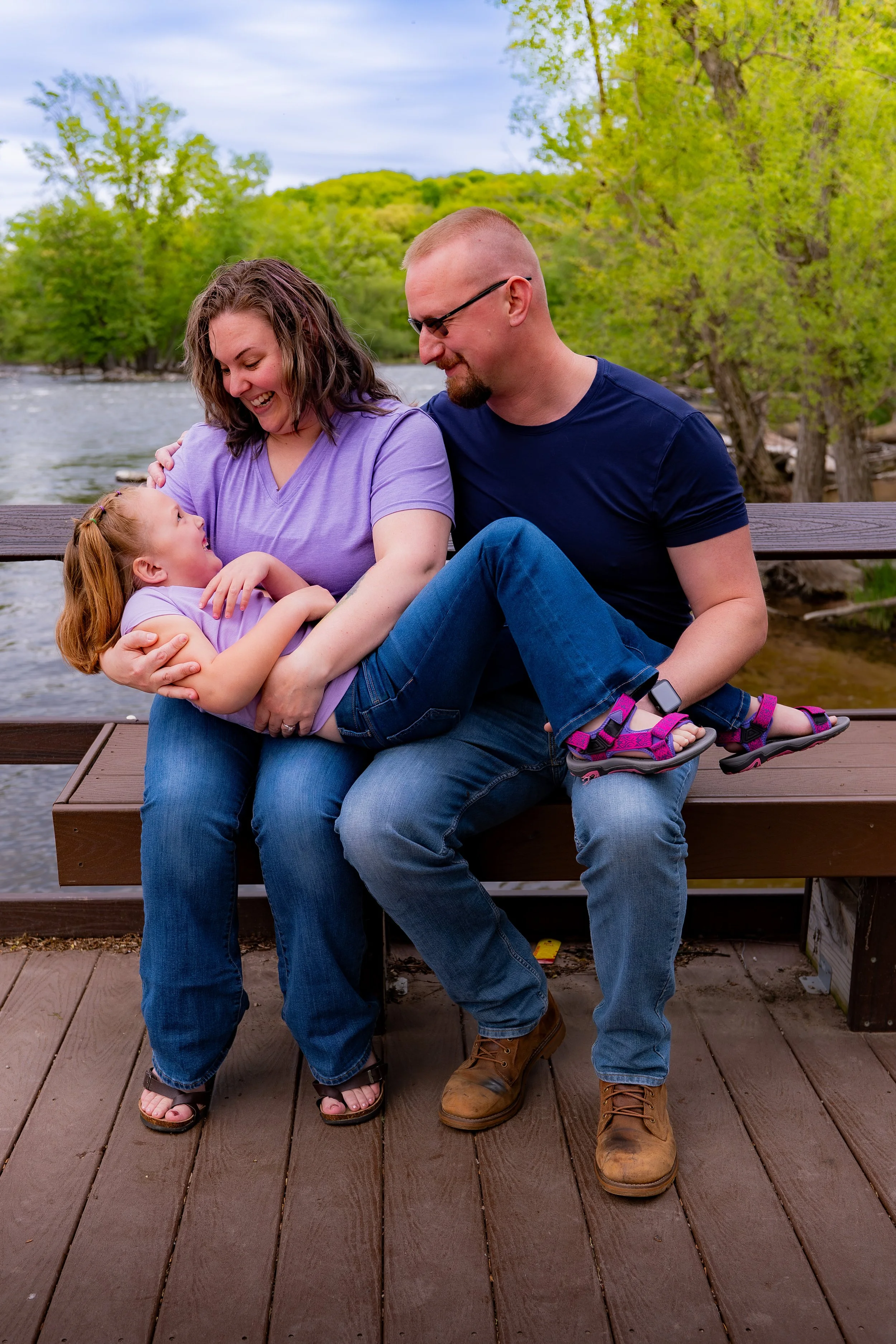 A family of three sitting together on a wooden dock by a river, surrounded by green trees and hills, with a cloudy sky overhead. The mother, father, and young daughter are smiling and engaged in a joyful moment.
