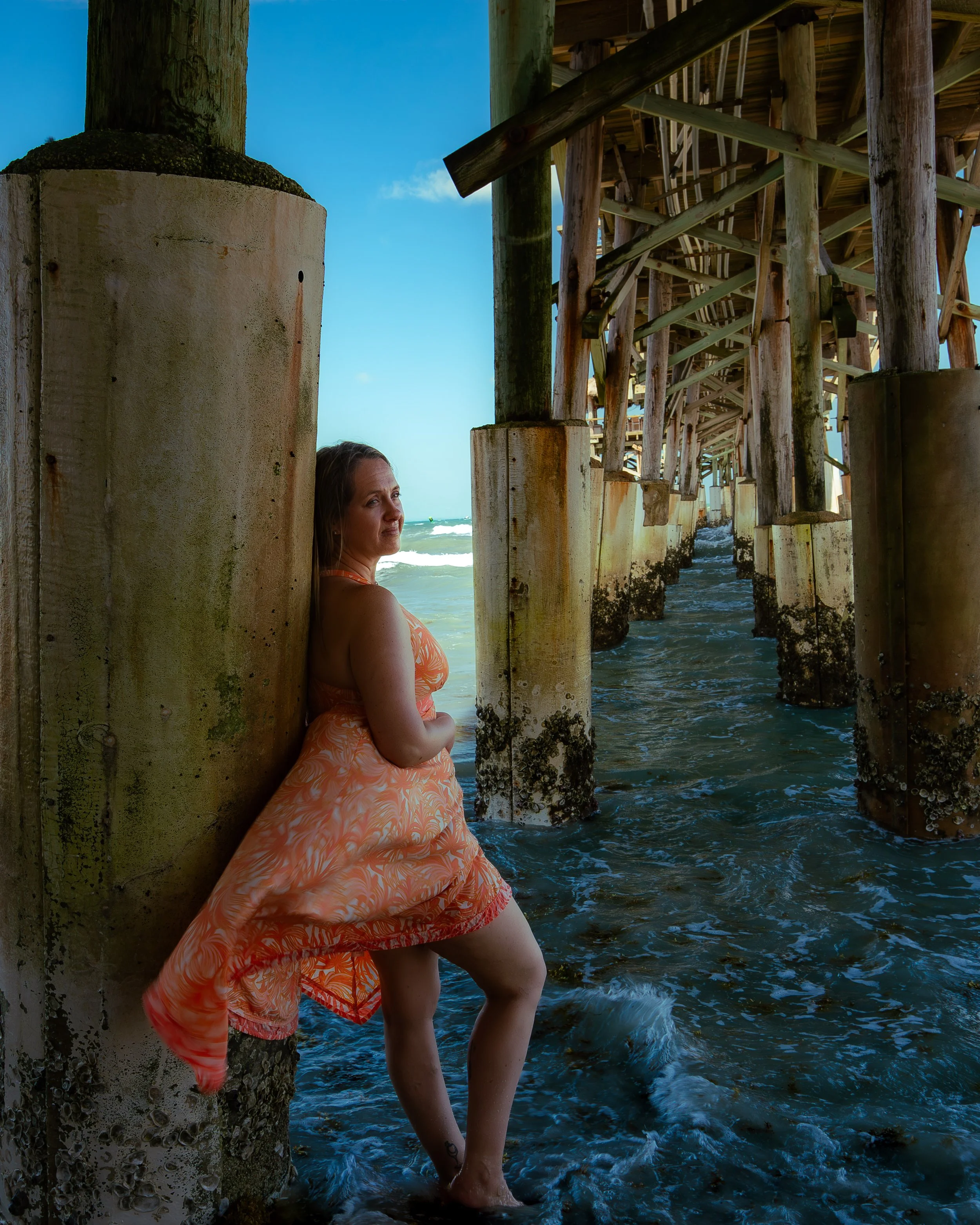 Woman in orange patterned dress standing in ocean water beneath a wooden pier on a sunny day.
