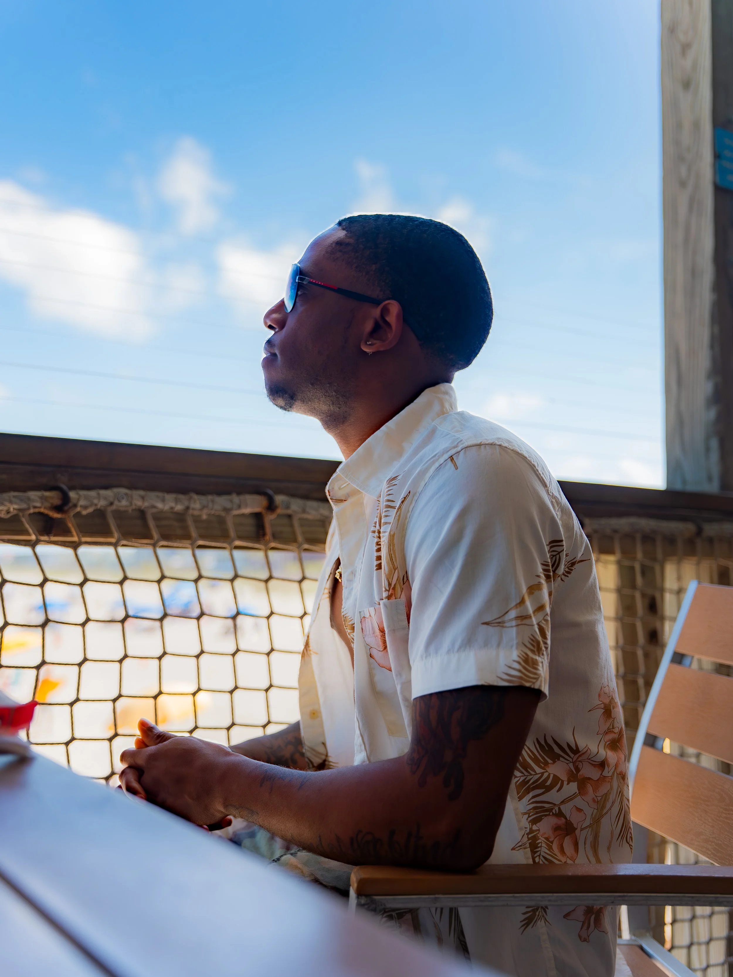 A man wearing sunglasses and a floral short-sleeve shirt is sitting outdoors at a table under a wooden roof structure, with a blue sky and some clouds in the background.