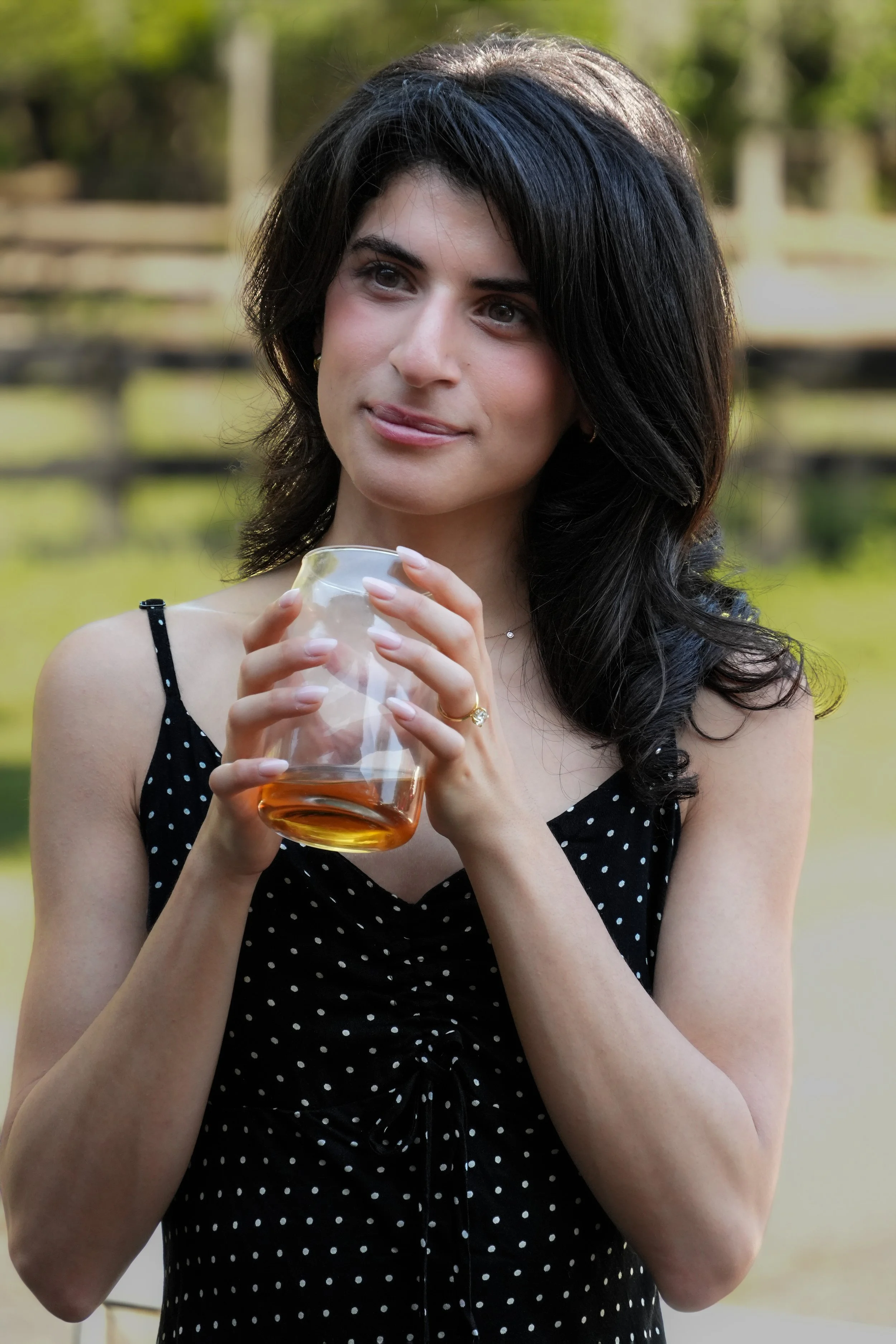 A young woman with dark, wavy hair holding a glass of amber-colored beverage outdoors, wearing a black dress with white polka dots.